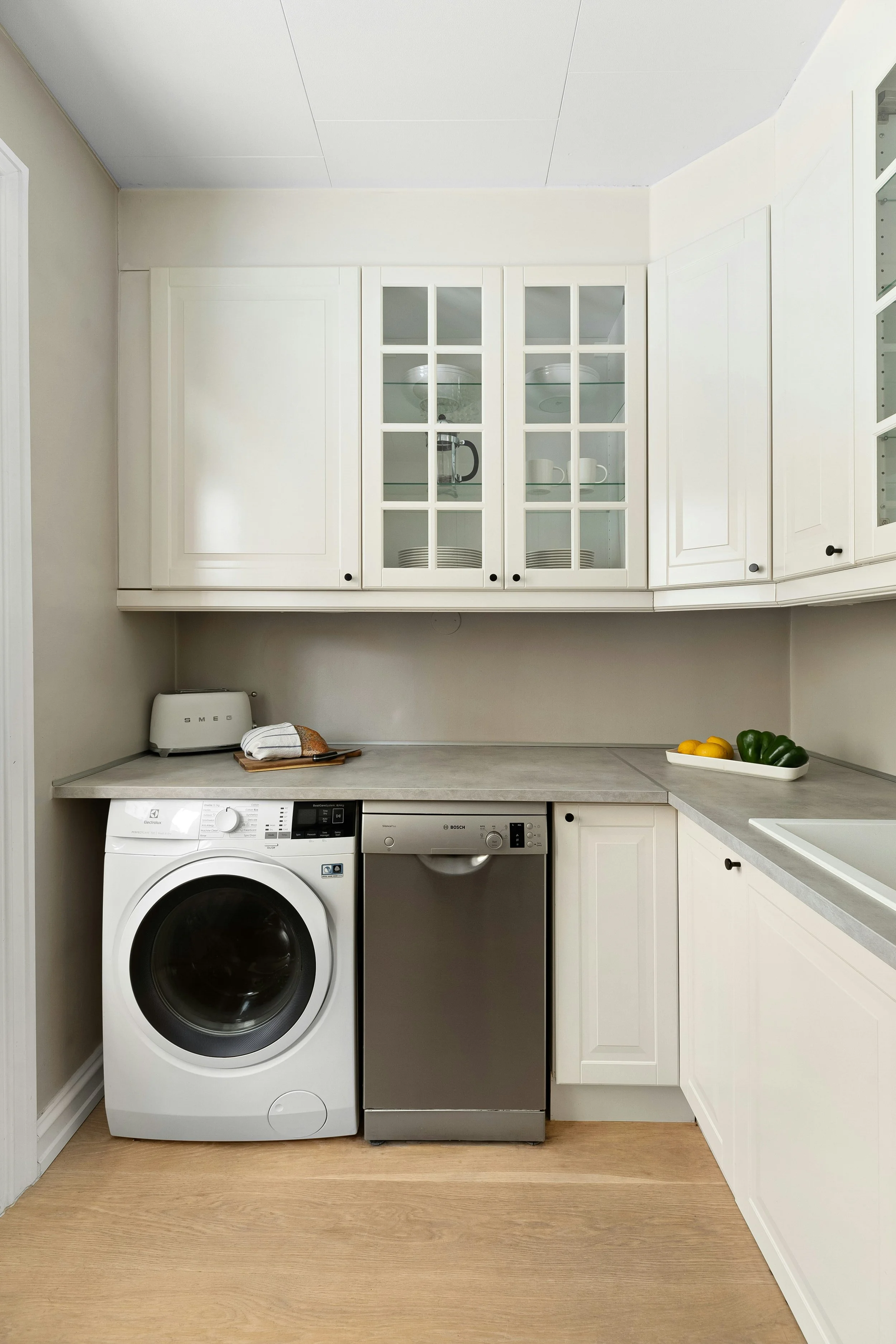 A kitchen laundry area with white cabinets, a washing machine, a dishwasher, and a countertop. Items on the countertop include a SMEG bread box, a tray with a towel, and a bowl of lemons and green bell peppers. The upper cabinets have glass doors with dishes inside.
