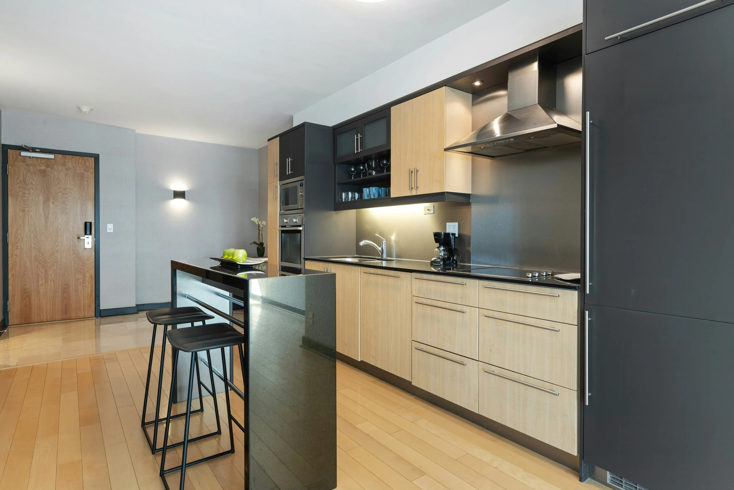 Modern kitchen with light wood cabinets, black accents, stainless steel appliances, and a black kitchen island with three black stools. There are apples and a plant on the island, and a door on the left side.