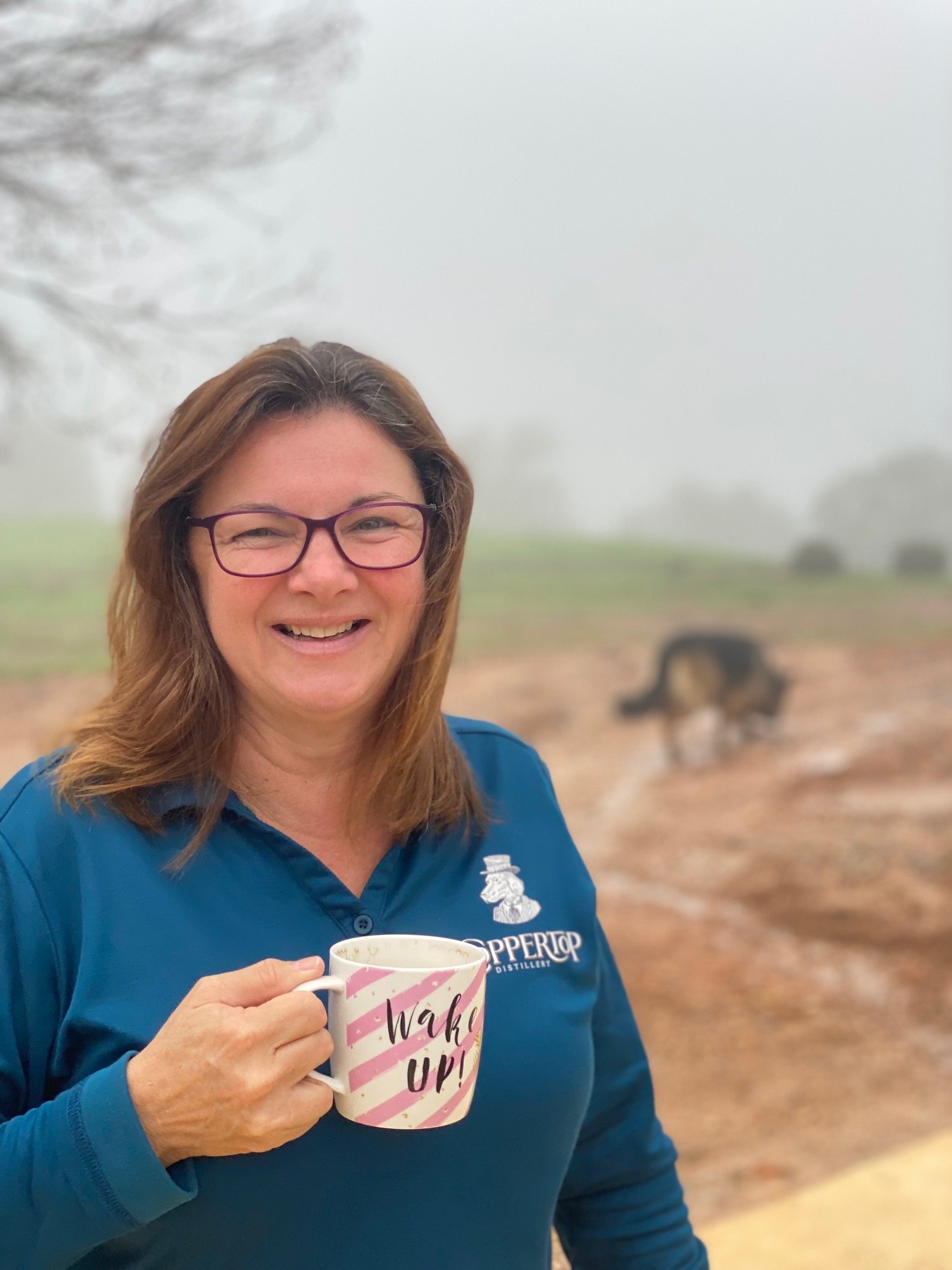 A woman smiling outdoors holding a mug that says 'Wake Up!'. She is wearing glasses and a blue jacket with a logo and text. There is a dog in the background on a foggy day.