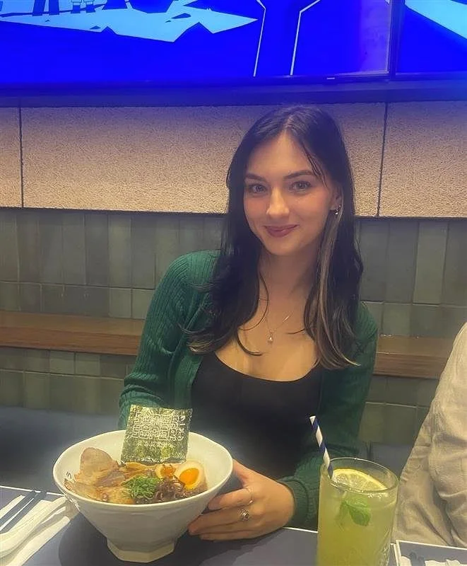 A young woman sitting at a table in a restaurant, holding a bowl of ramen with various toppings and a QR code on a piece of seaweed. She has long dark hair, is wearing a black top and green cardigan, and is smiling at the camera. Next to her is a yellow drink with a straw and mint leaves, garnished with lemon.
