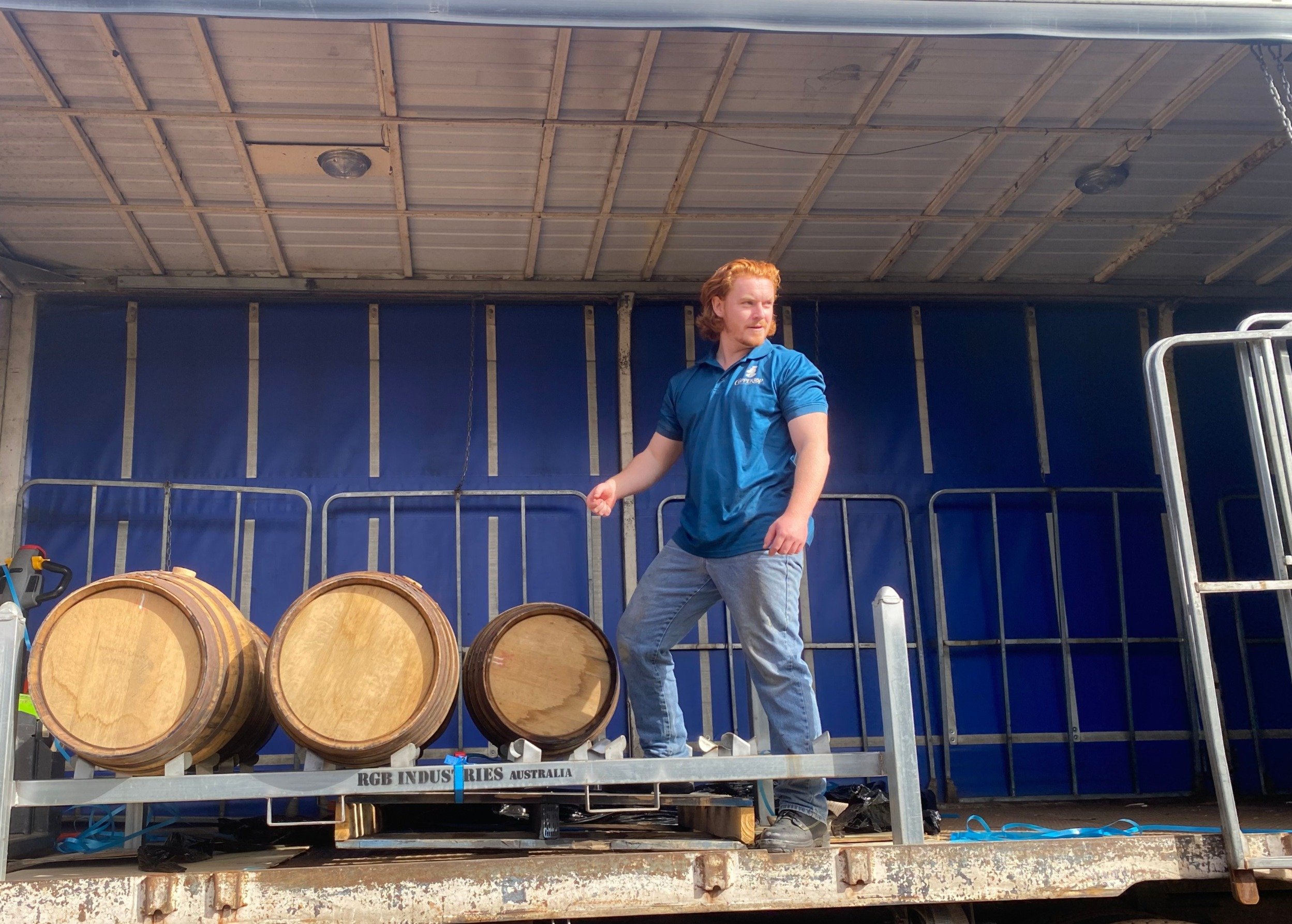 A man in a blue shirt and jeans standing on a flatbed truck, with three large wooden barrels in front of him, inside a garage or warehouse with a blue background.
