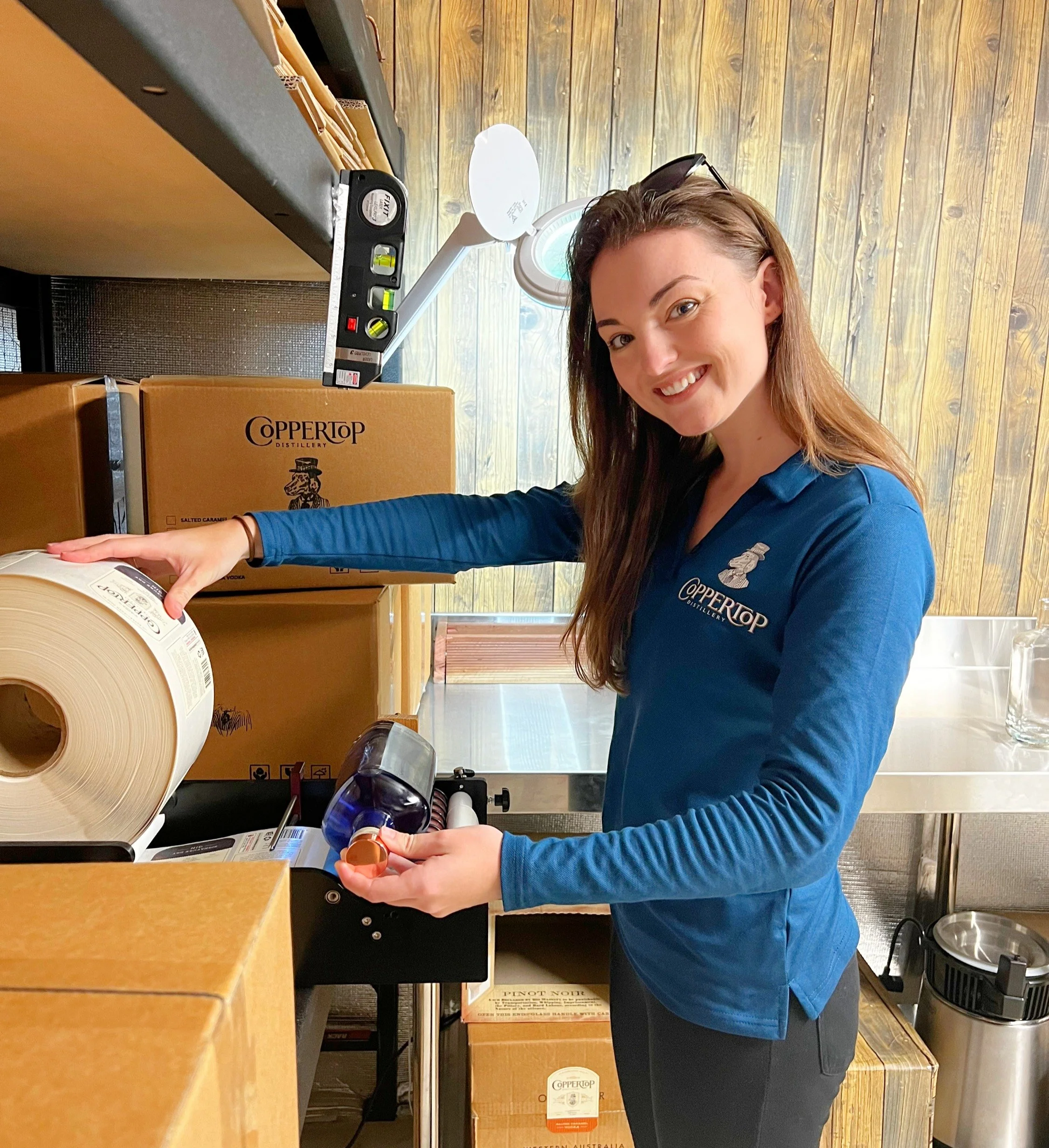 Woman with long brown hair smiling, wearing a blue jacket with the "Coppertop" logo, handling a spool of paper or tape in a store with cardboard boxes and cleaning supplies nearby.
