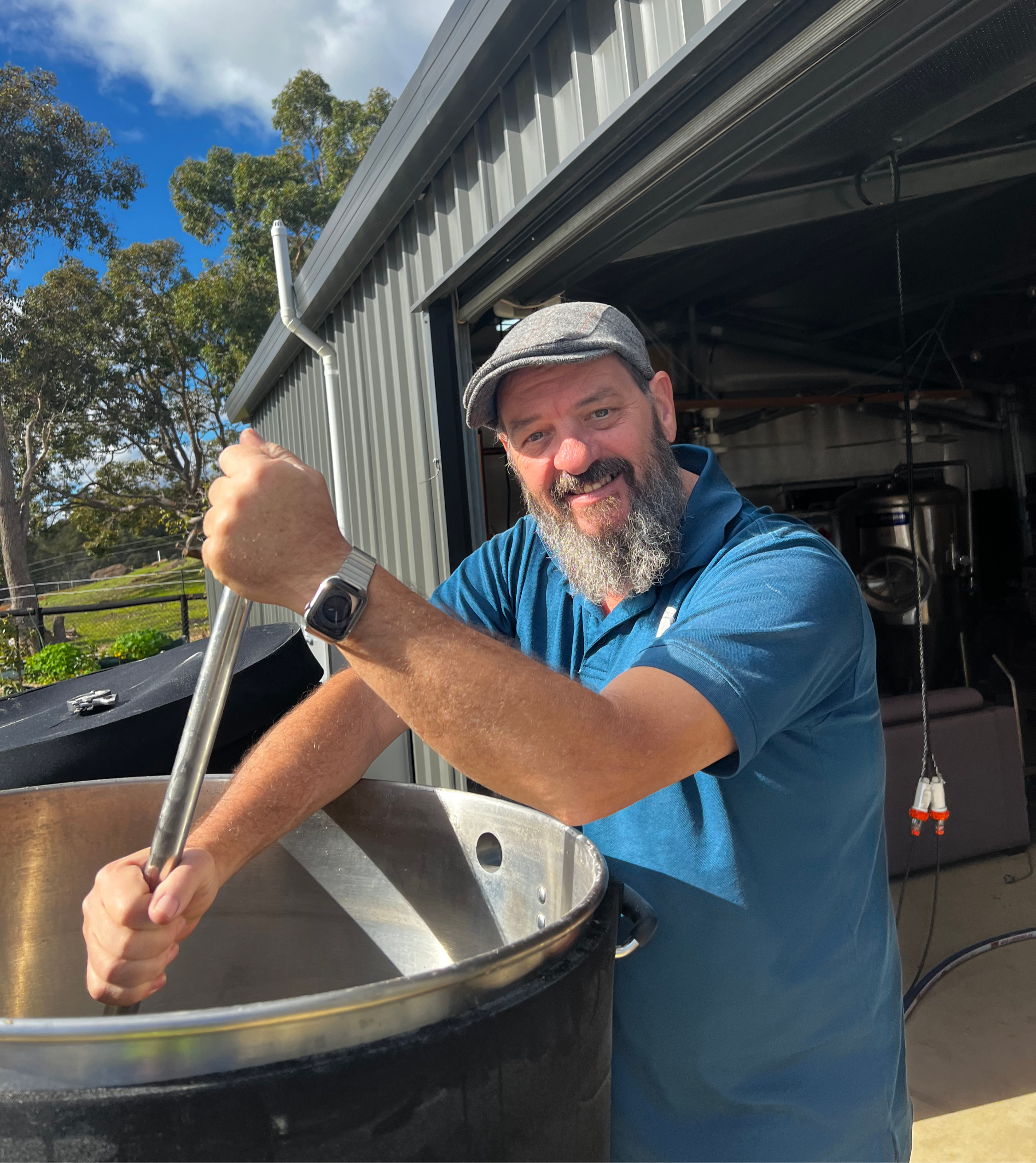 A man with a gray beard wearing a gray cap and blue shirt stirring a large cooking pot outdoors on a sunny day.
