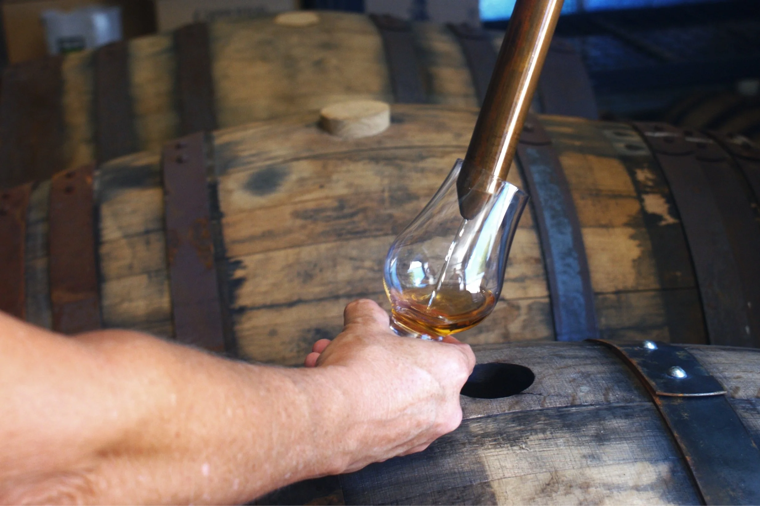 A person pours bourbon whiskey from a copper still into a glass inside a distillery with wooden barrels in the background.