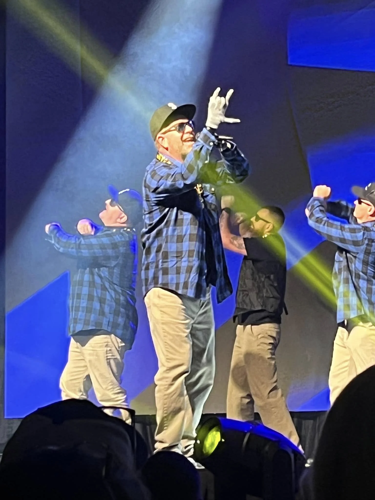 Man performing on stage with backup dancers, wearing glasses, a black cap, a checkered shirt, and khaki pants, illuminated by stage lights.