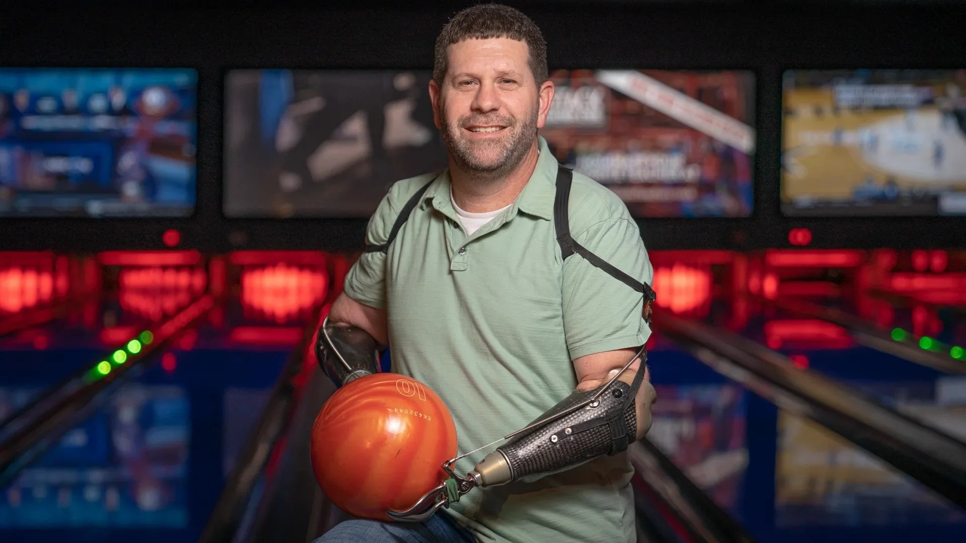 Man with a prosthetic arm holding an orange bowling ball at a bowling alley.