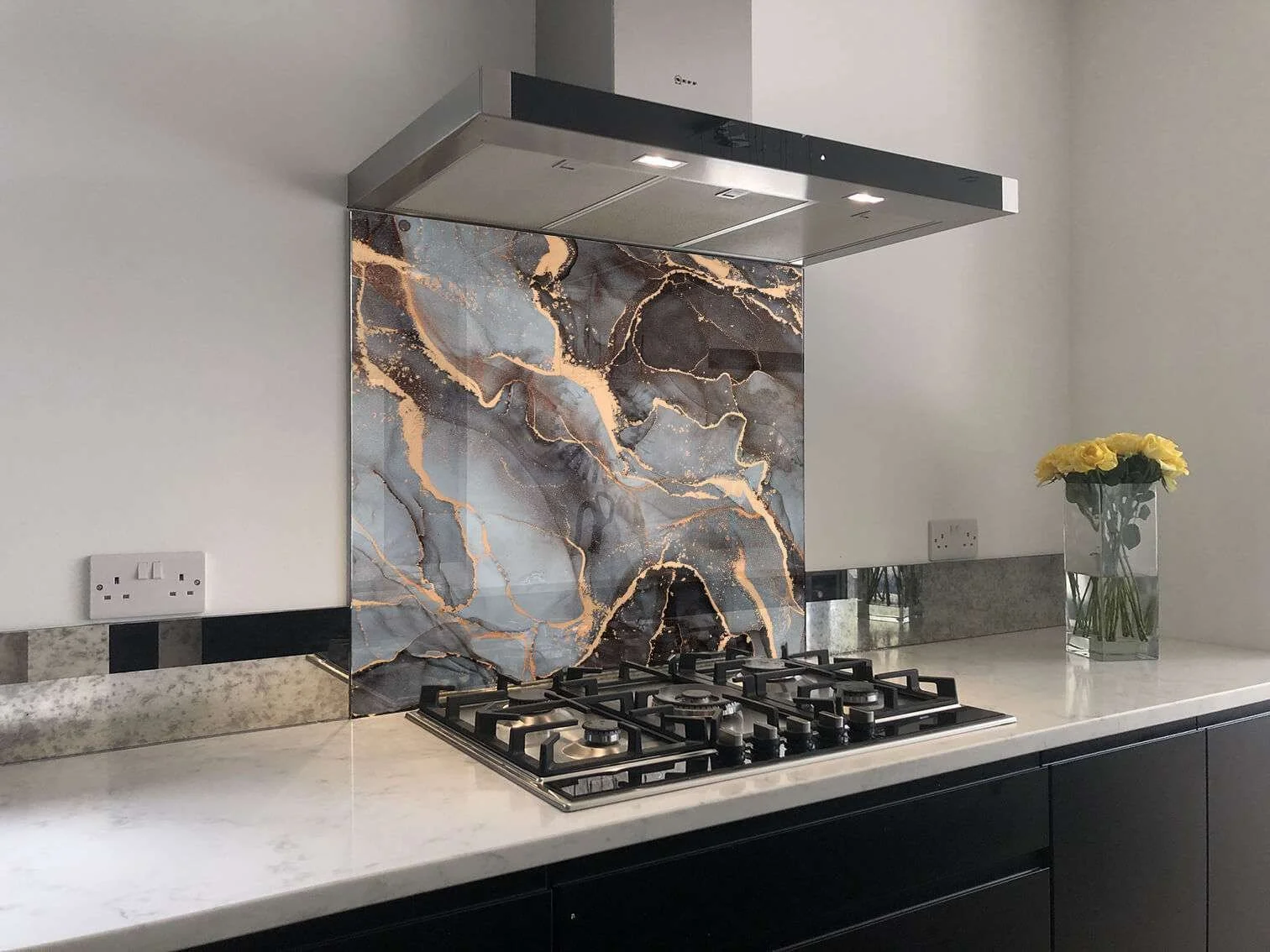 Modern kitchen countertop with black gas stove, marble backsplash with blue, black, and copper veining, and a vase of yellow flowers on the right side.