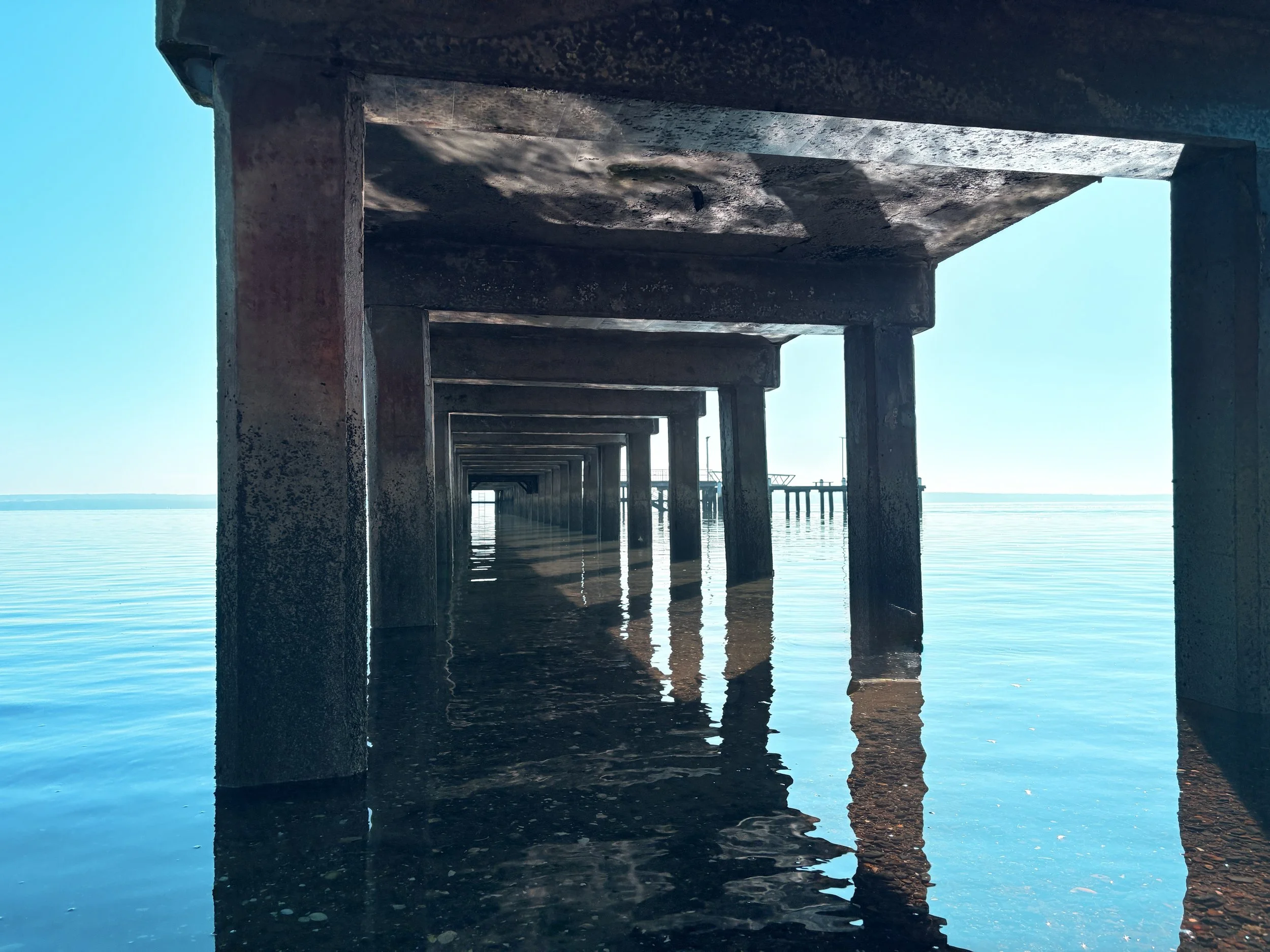 Underwater view of a concrete pier extending into a calm body of water, with blue sky in the background.