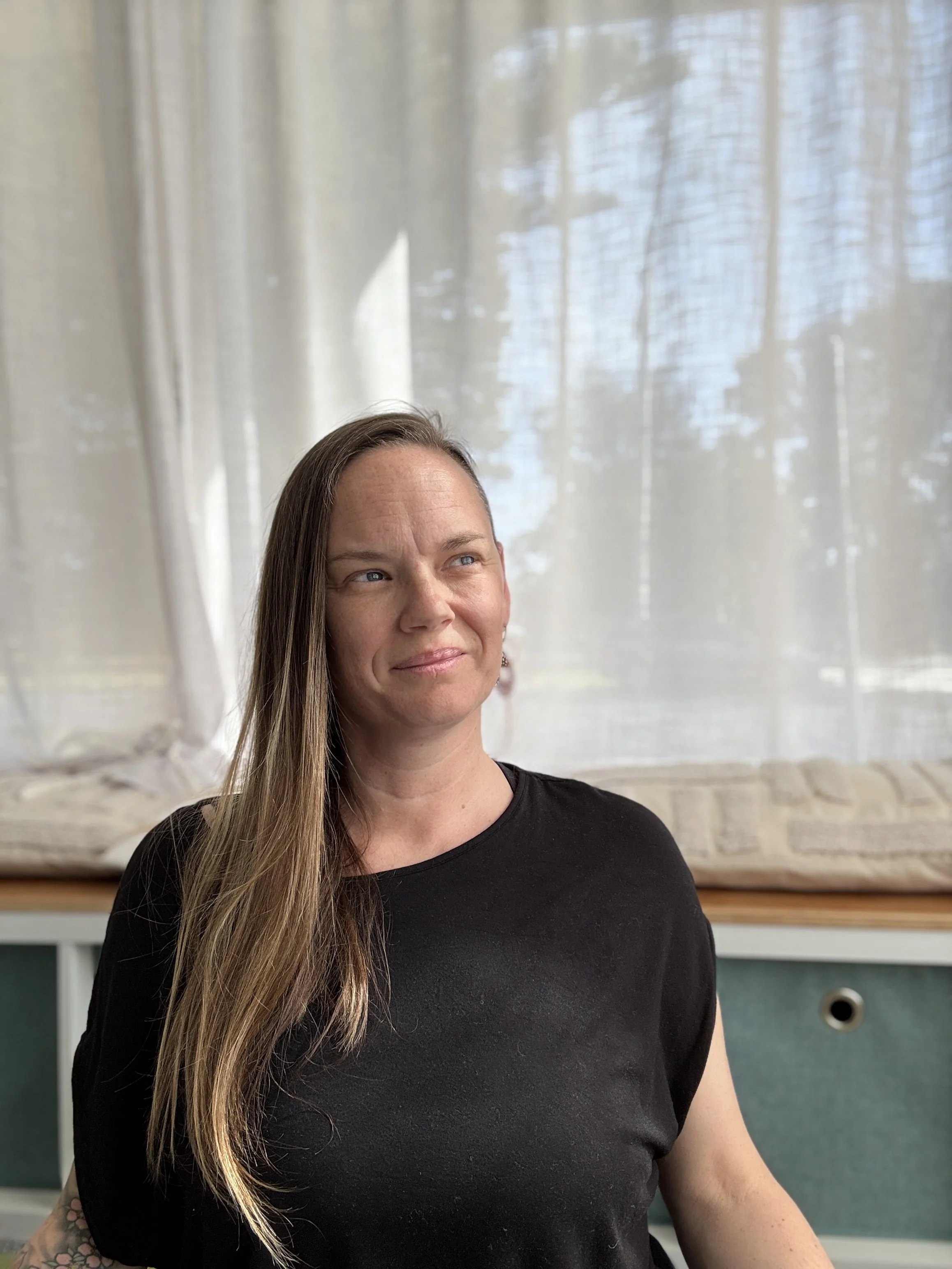 A woman with long hair wearing a black shirt sitting indoors with a light-colored curtain and window in the background.