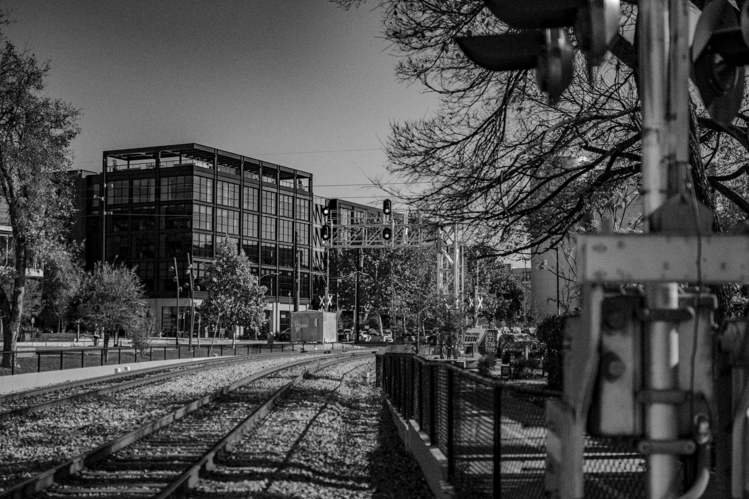 City street scene in black and white with railway tracks in foreground, modern multi-story glass building, traffic lights, and trees lining the street. Austin Texas.