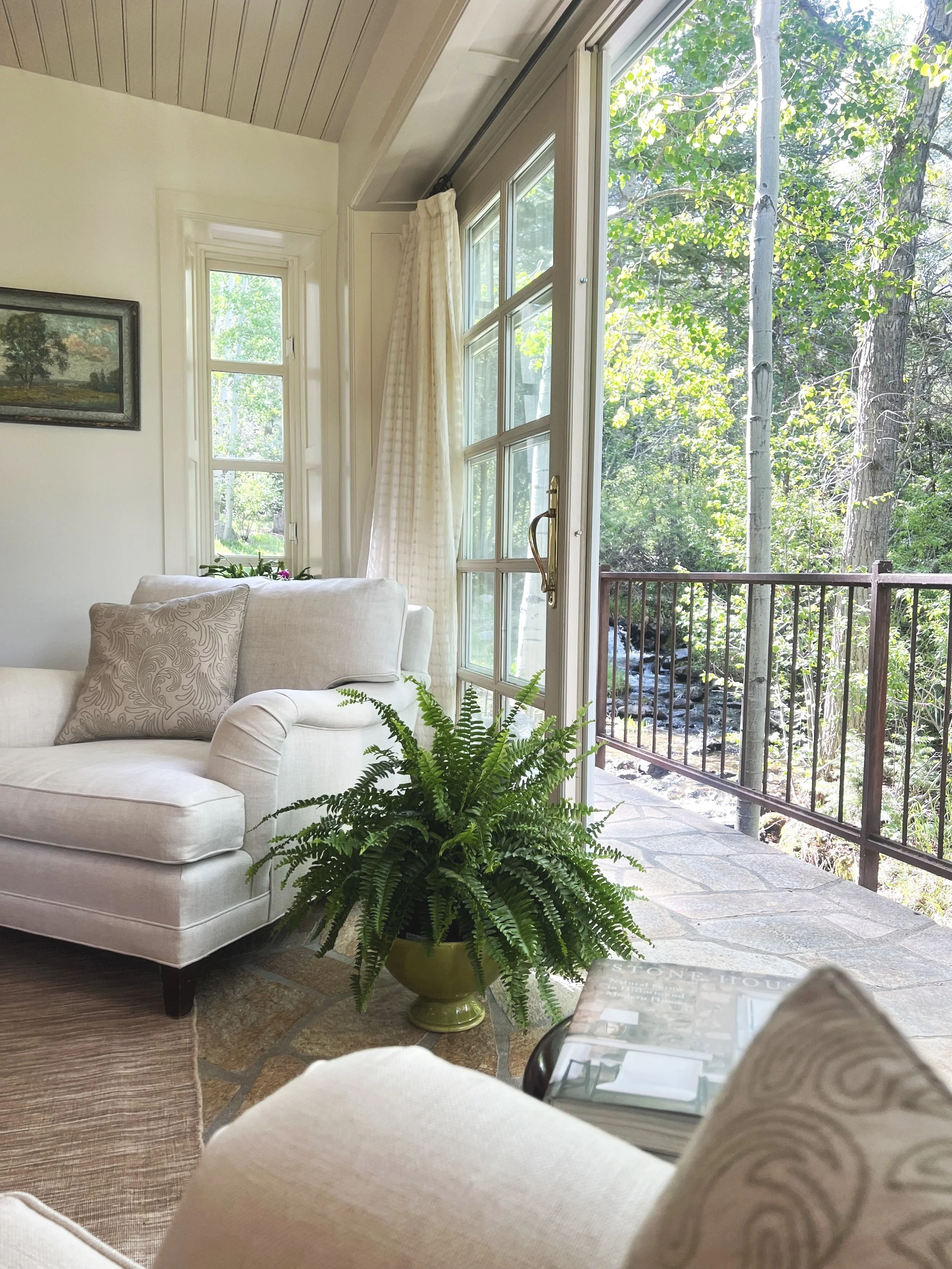 Living room with white sofas, a green potted fern, and large windows showing a view of trees and a creek outside.