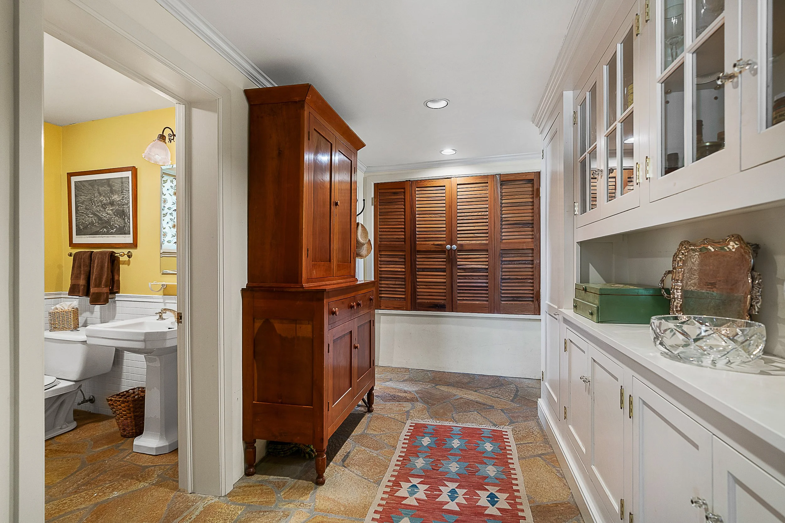 View of a bright, cozy hallway with wooden cabinetry and a window with wooden shutters, leading to a bathroom with yellow painted walls and a framed picture.