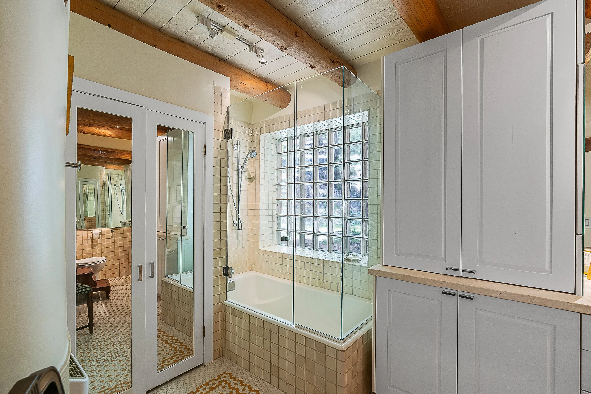 Bathroom with a bathtub enclosed by glass, a window with glass blocks, a white cabinet, and a door with a mirror leading to a toilet area.