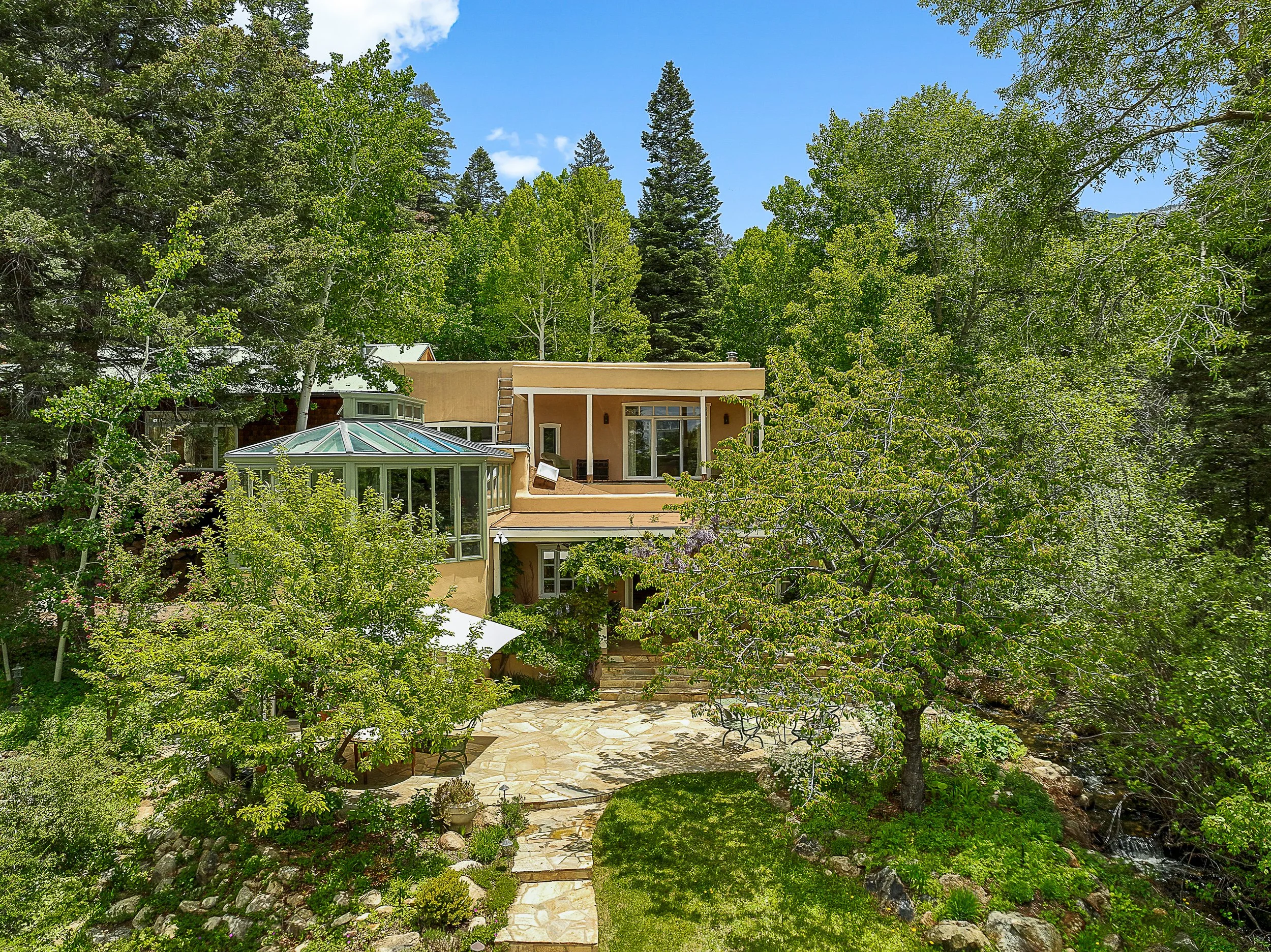 A multi-story house with beige exterior walls, large windows, and a glass-enclosed sunroom, surrounded by lush green trees and a landscaped yard with stone pathways and a small waterfall.