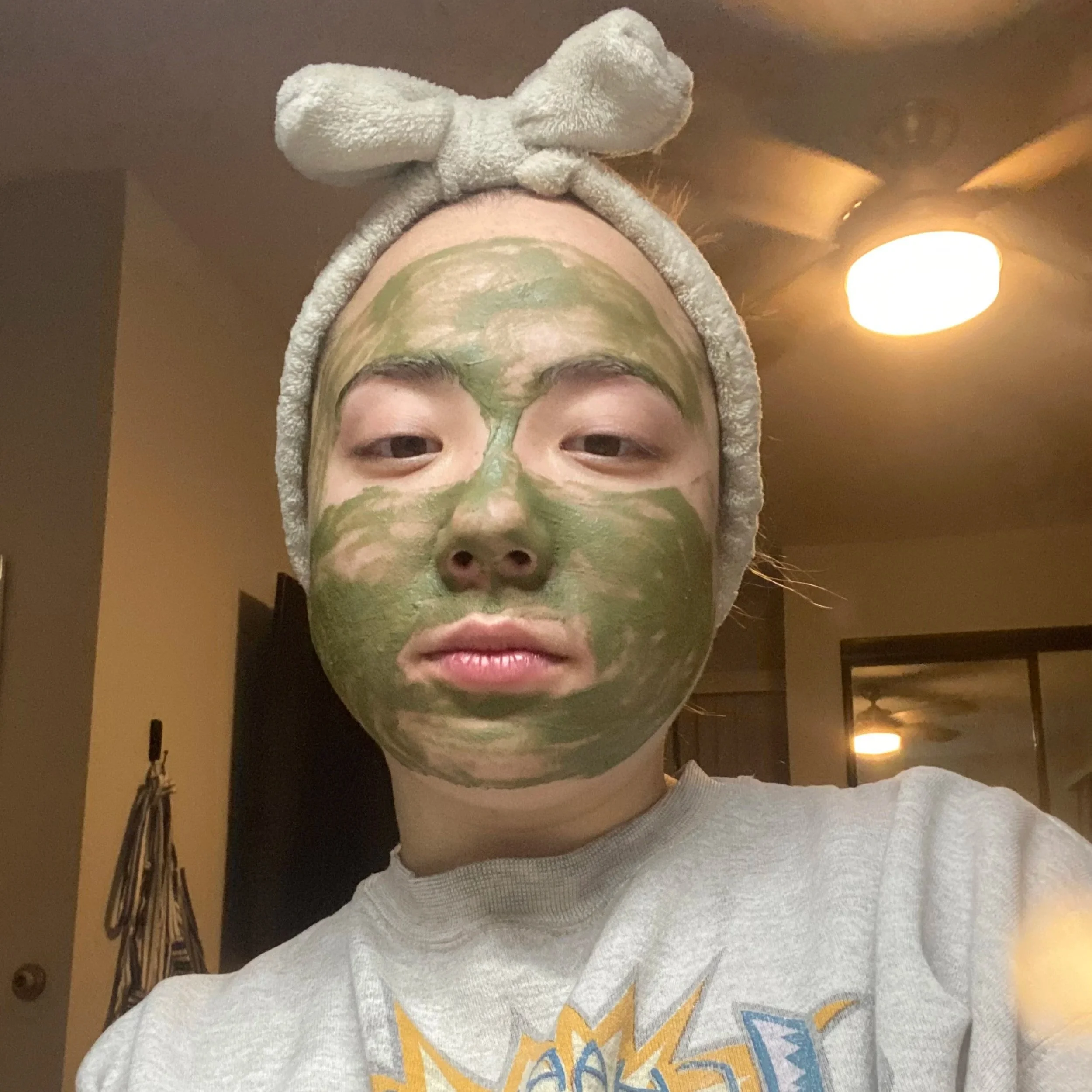 Person with a green facial mask wearing a beige headband with a bow, indoors with ceiling lights and a mirror in the background.