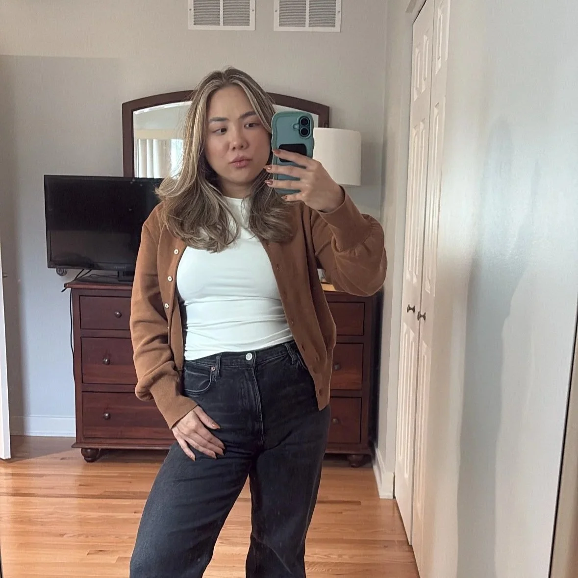 A woman taking a selfie in a bedroom mirror, wearing a brown jacket, white t-shirt, and black jeans, with a dresser, TV, and white door in the background.