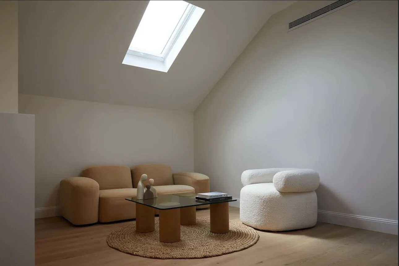 Minimalist living room with beige sofa, white textured lounge chair, glass coffee table, small decorative sculptures, magazines, round woven rug, under a sloped ceiling with a skylight and air vent.