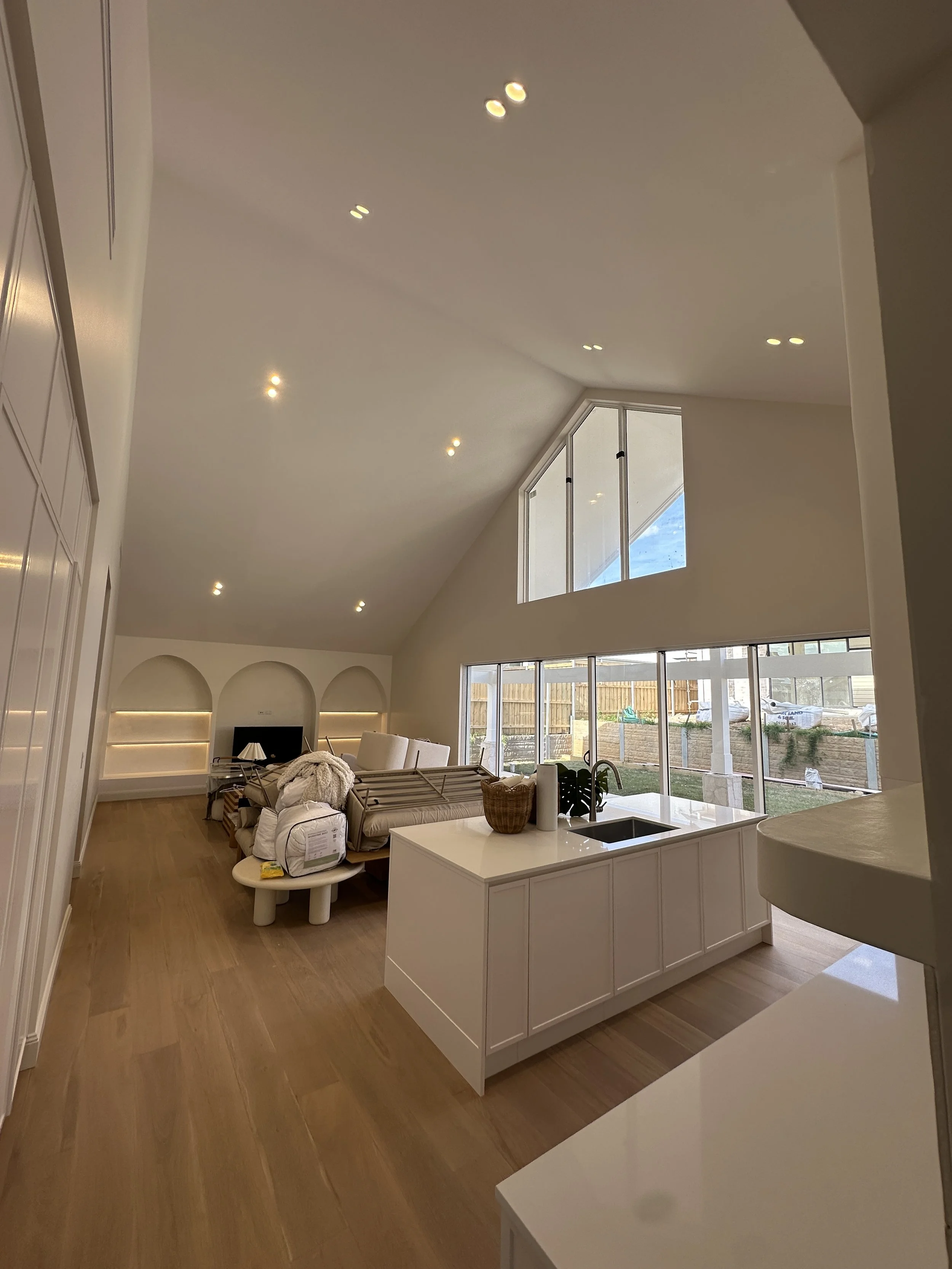 Interior view of a modern living room with high vaulted ceiling, large windows, and light wood flooring. There are built-in shelves with accent lighting, a white kitchen island with a sink, and furniture covered with protective wrapping, indicating ongoing setup or renovation.