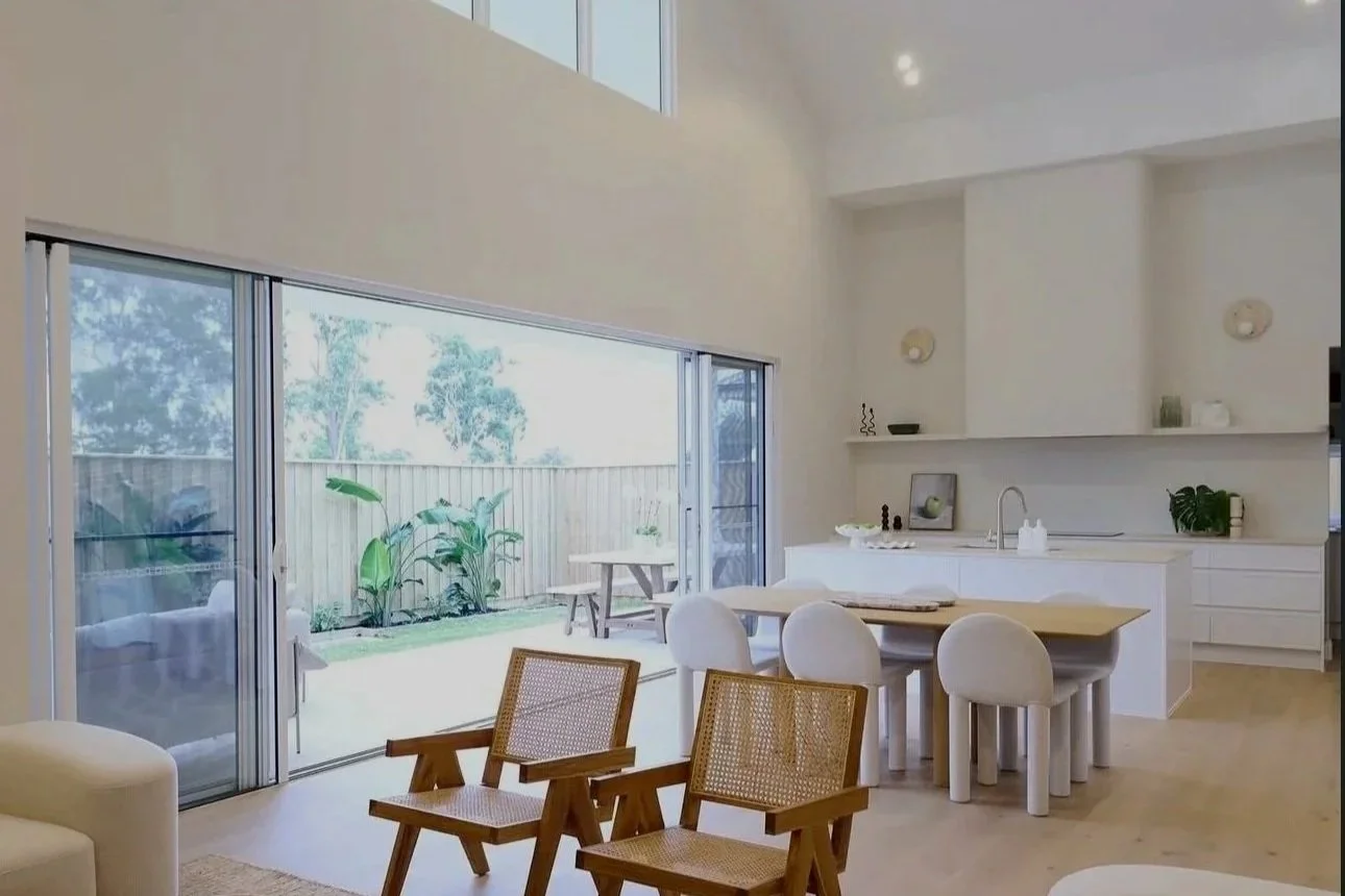 Bright living area with sliding glass doors leading outside, including a small patio with a picnic table, wooden chairs, and minimalist decor with white walls and built-in shelves.