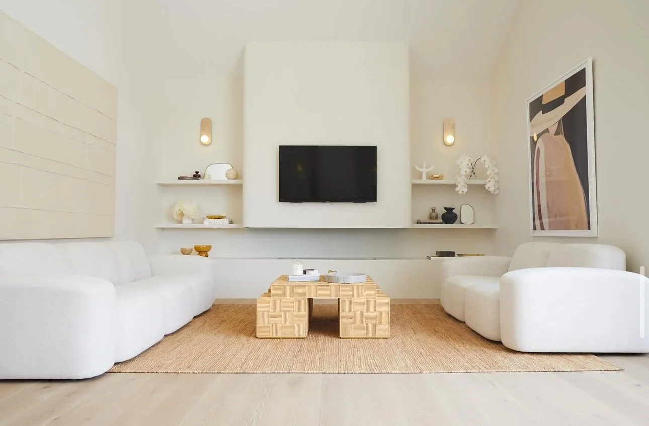 A modern living room with white sofas, a wooden coffee table, wall-mounted shelves with decorative objects, and a black flat-screen TV on a white wall.