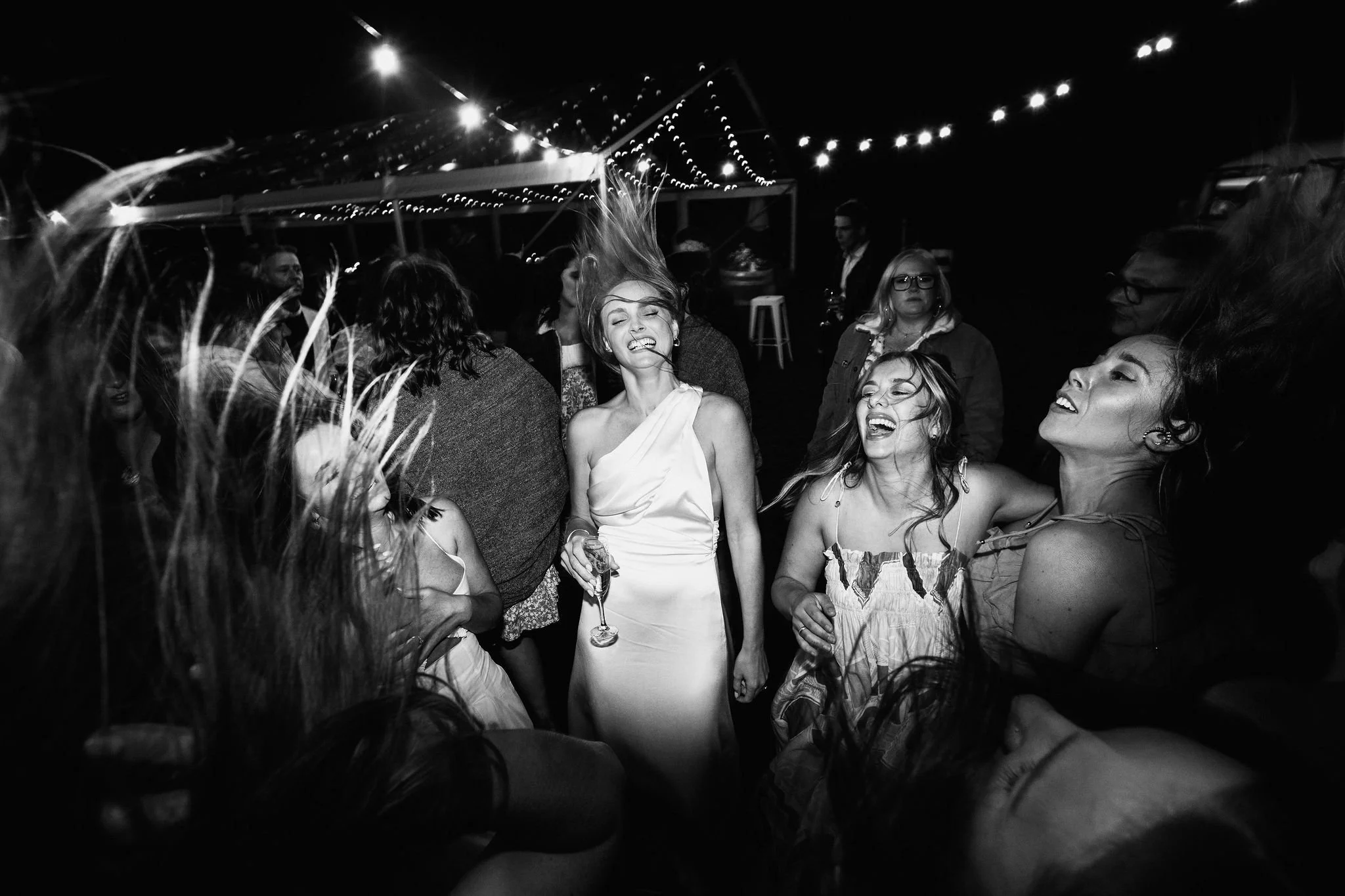 Group of people dancing and enjoying themselves at a party or celebration, with string lights overhead, in black and white.