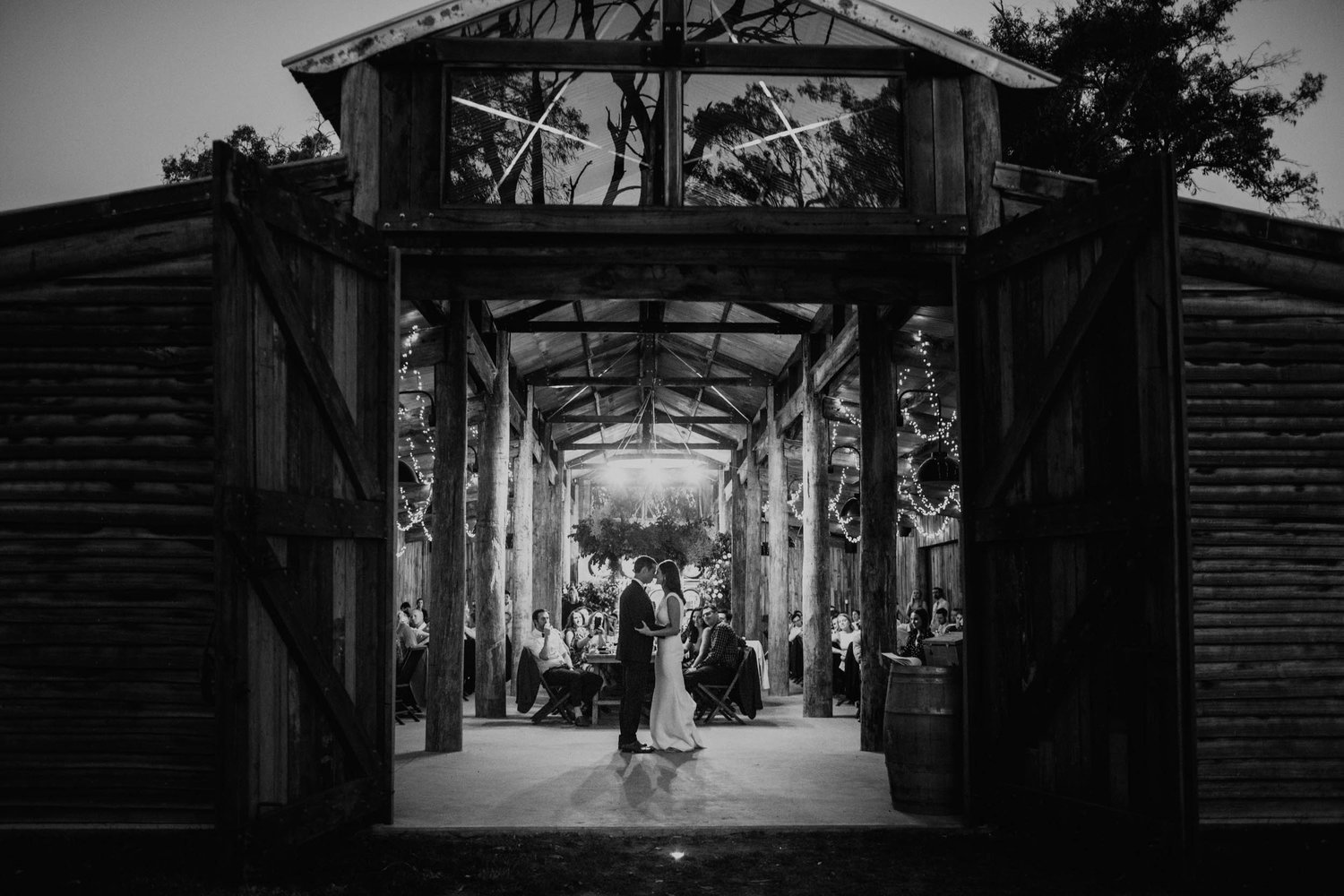 A black and white photo of a barn wedding reception with a couple dancing in the center and guests sitting at tables around them. Open barn doors frame the scene with string lights hanging inside and large windows at the top.