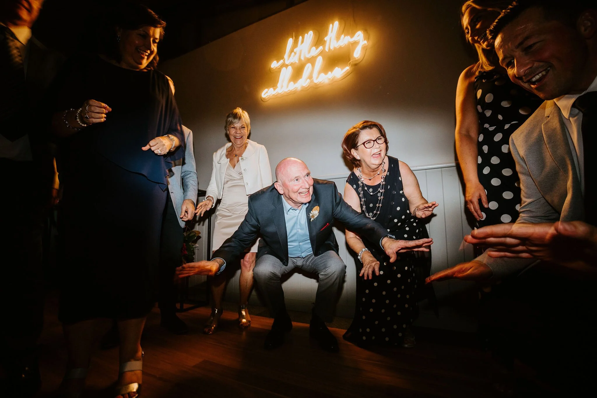 Group of elderly people dancing and celebrating at a party with a neon sign that reads 'a little thing called love' in the background.