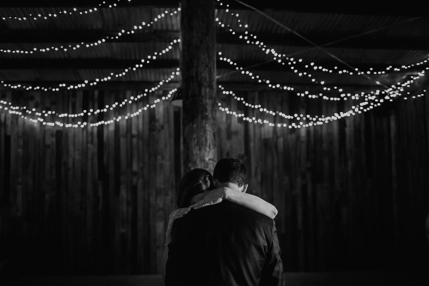 A black and white photo of a couple embracing, with the woman’s arms around the man's shoulders, in a rustic setting with string lights overhead.