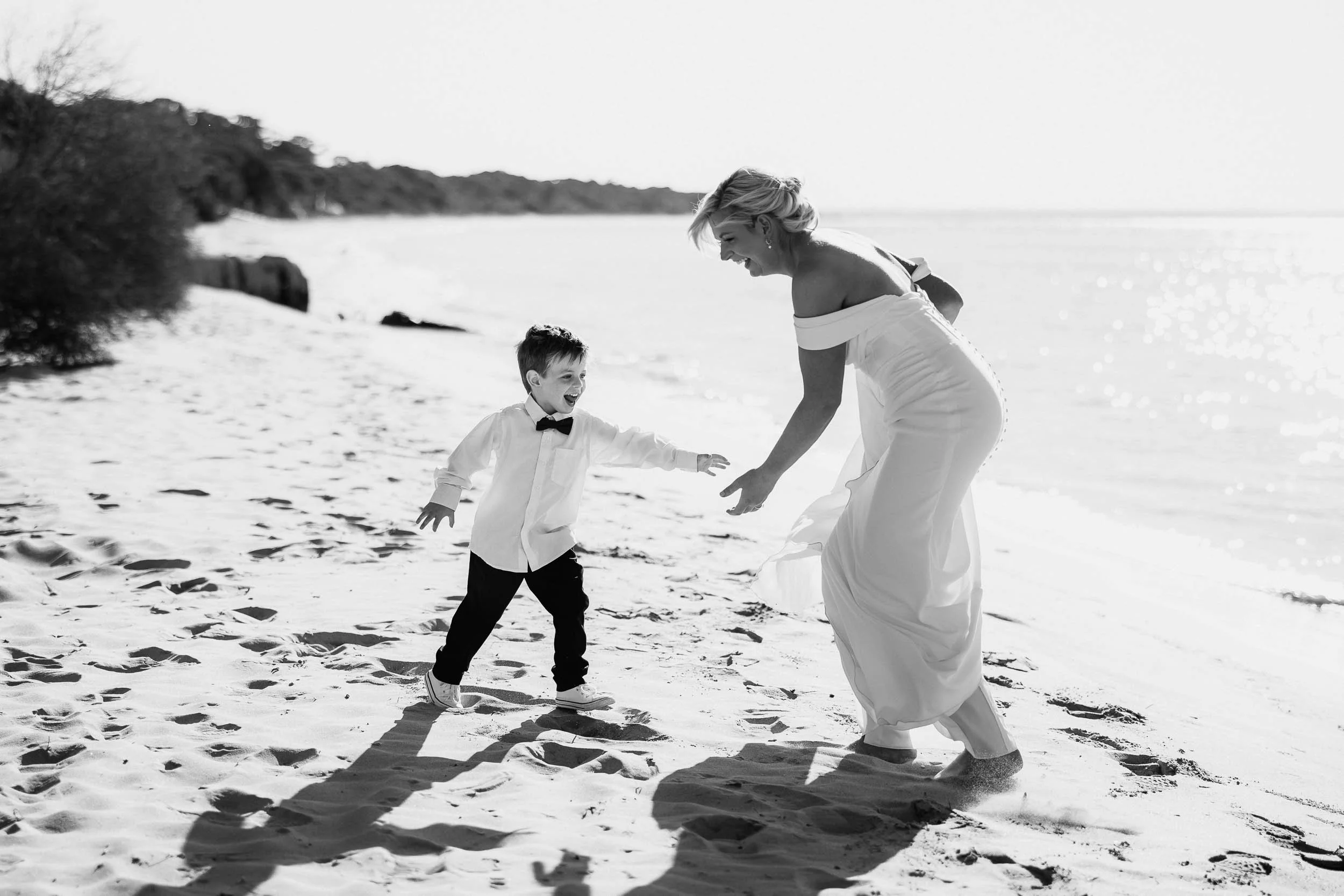 A woman and a young boy enjoying a playful moment on a sandy beach, both dressed in formal attire, with the woman in a gown and the boy in a shirt and bow tie, capturing a joyful scene.
