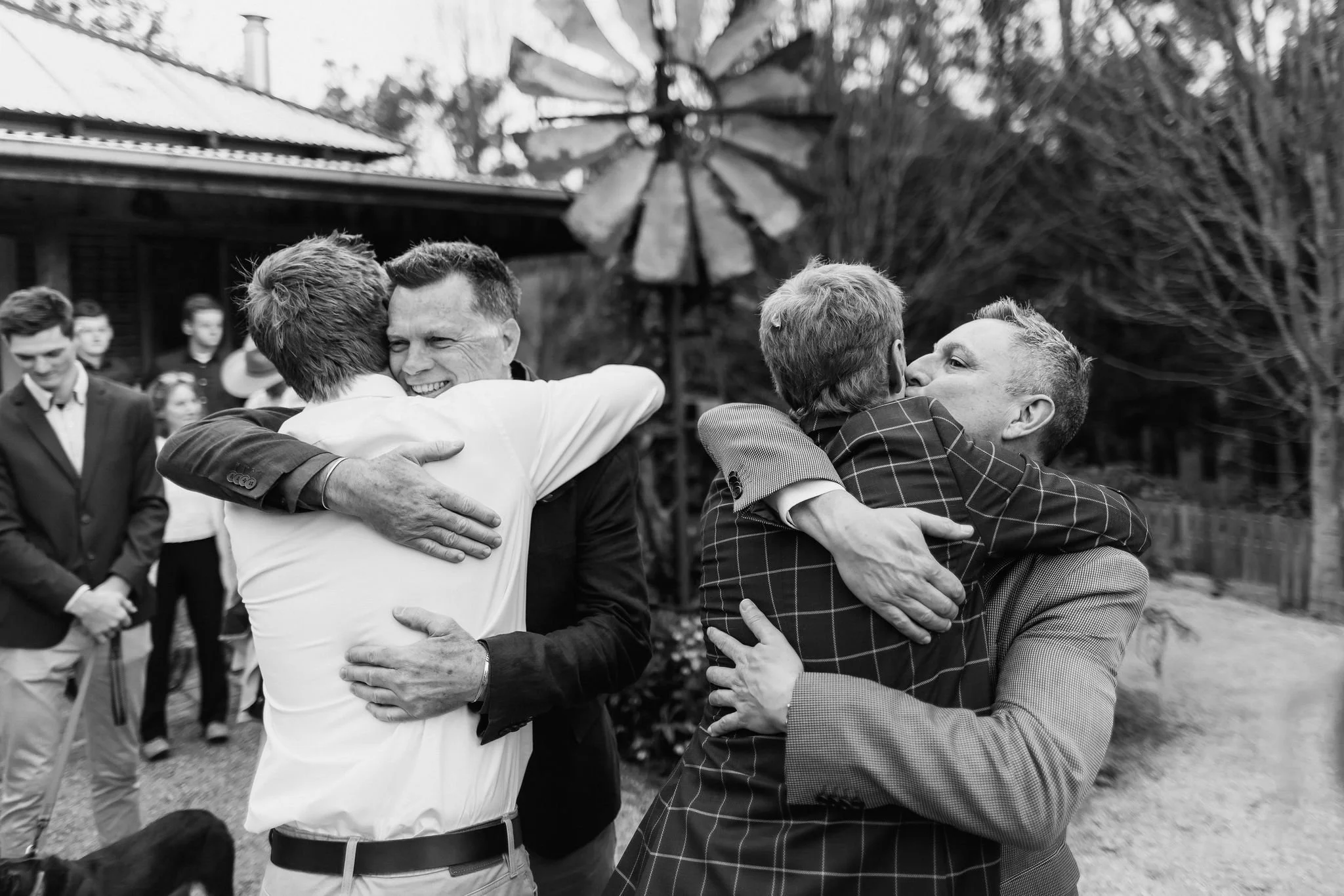 Two pairs of men hugging and smiling at an outdoor gathering, with a group of people watching in the background.