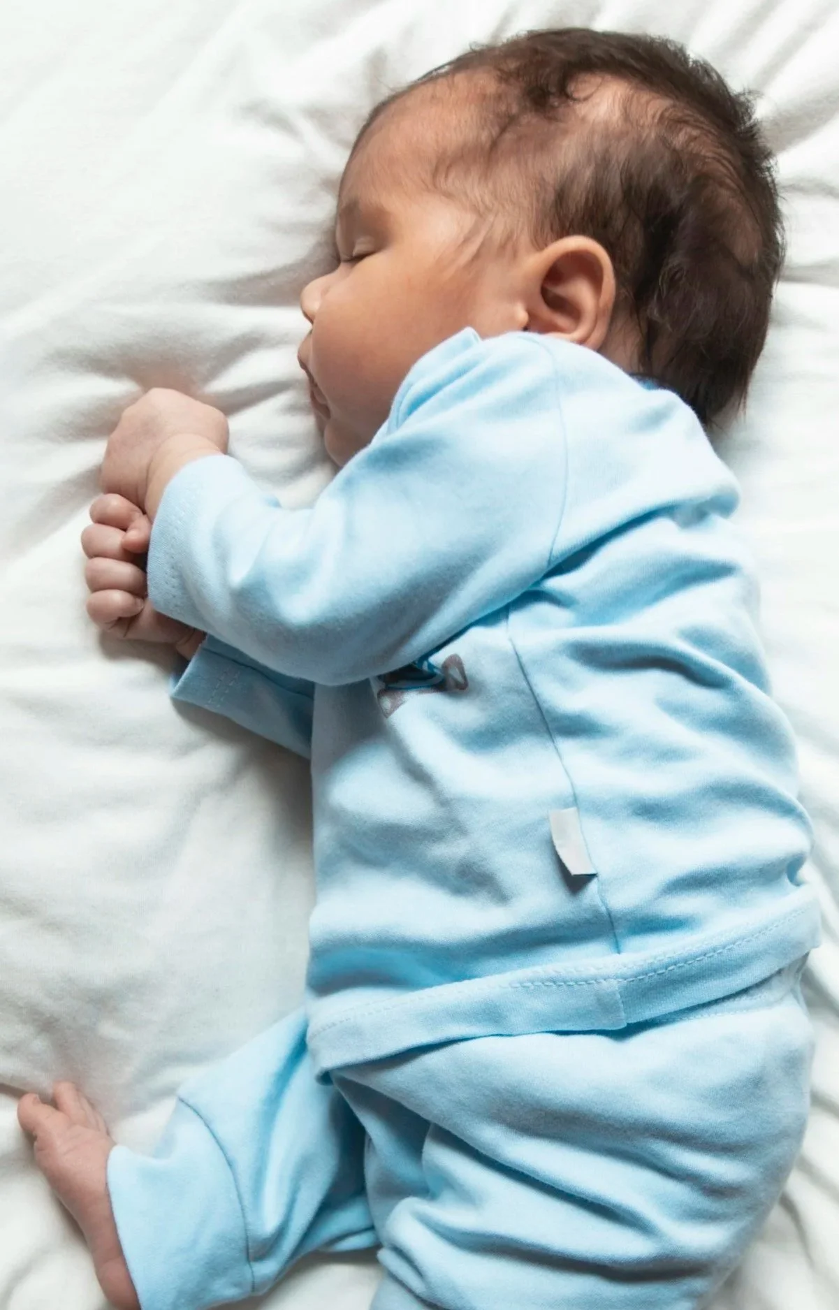 A sleeping baby dressed in light blue lying on a white blanket.