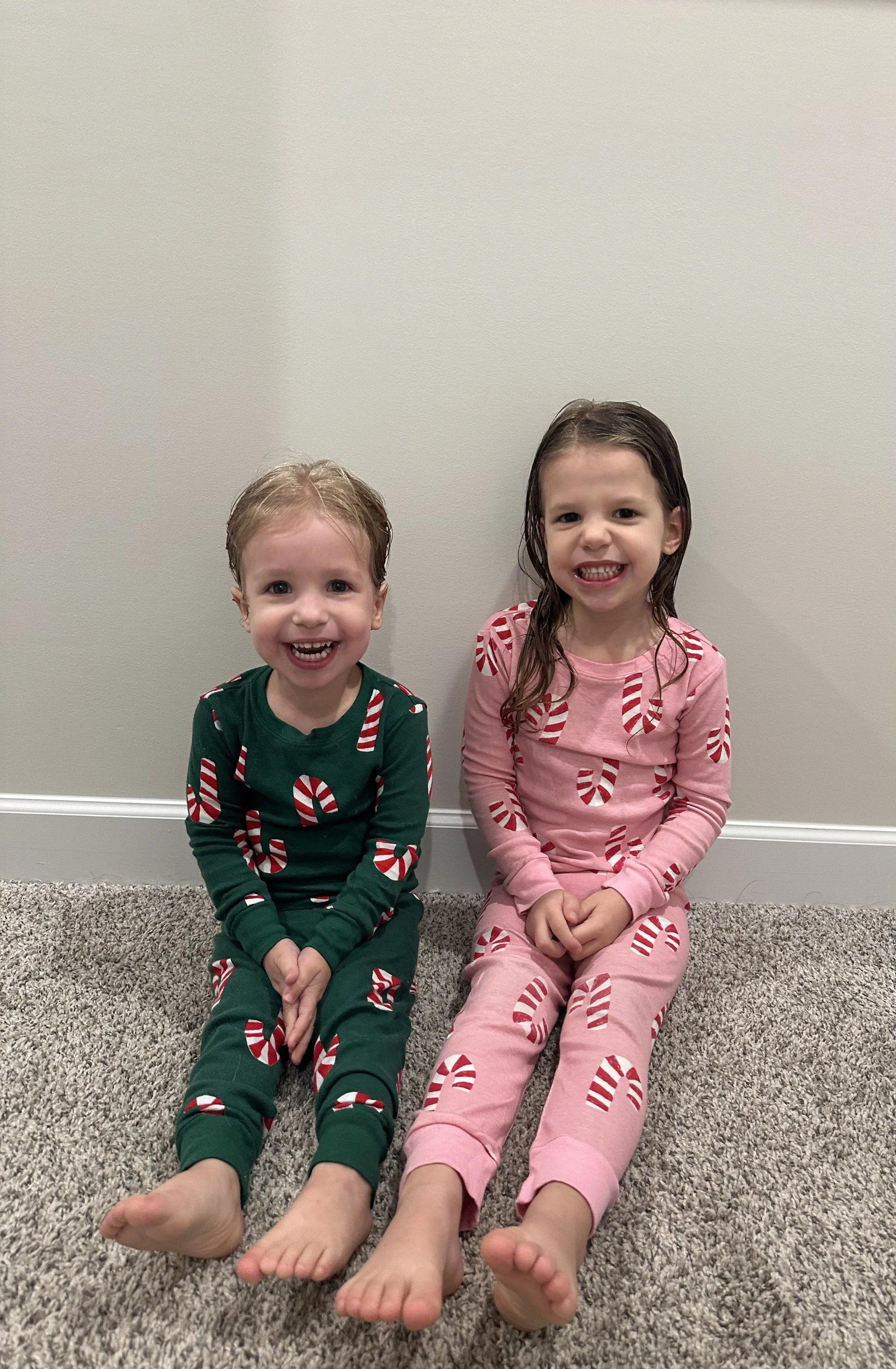 Two young children sitting on a carpeted floor, dressed in Christmas-themed pajamas with candy cane patterns, smiling happily at the camera.