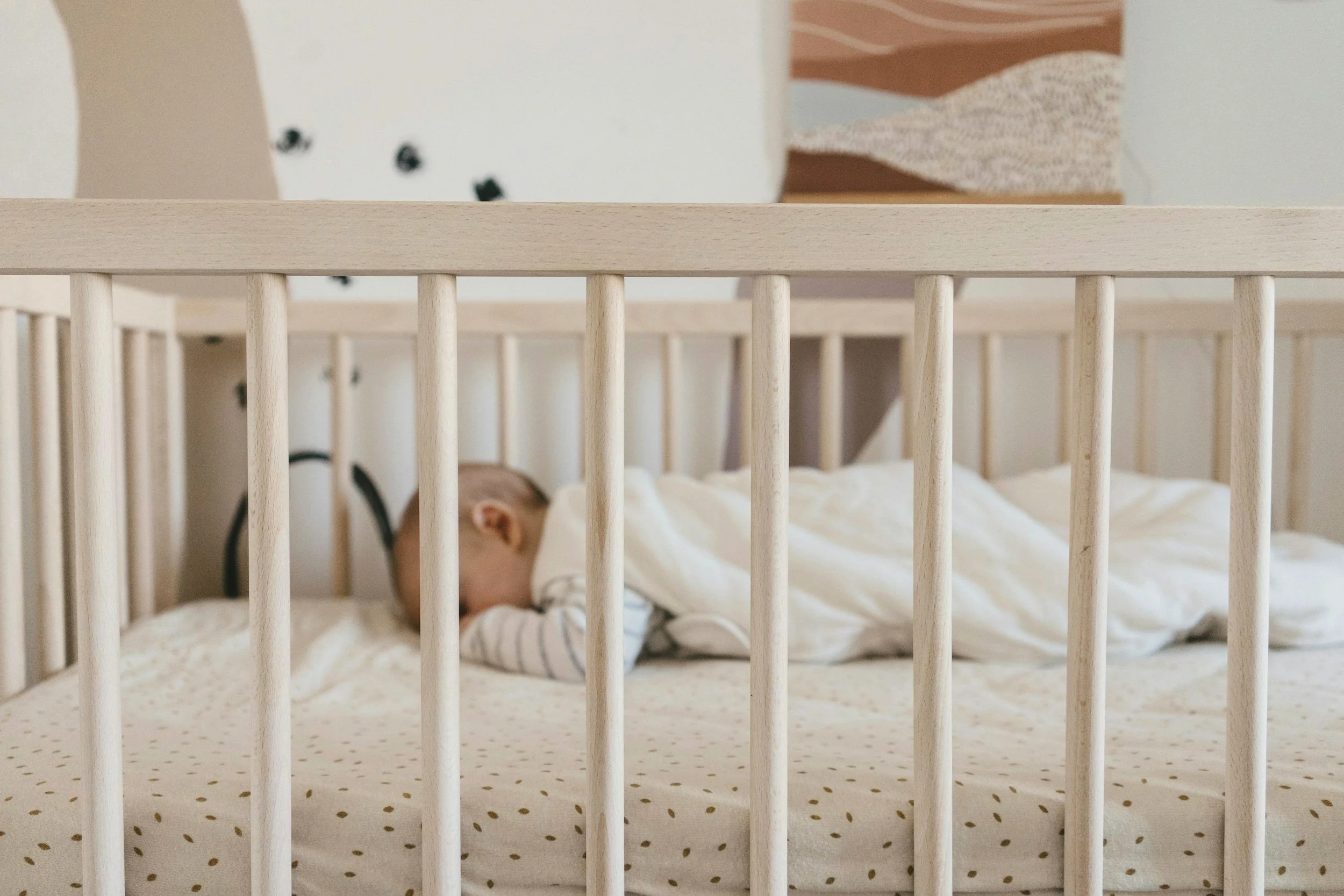 A baby sleeping in a crib with white wooden bars, dressed in a white onesie, in a nursery with light-colored walls and a piece of artwork hanging above.