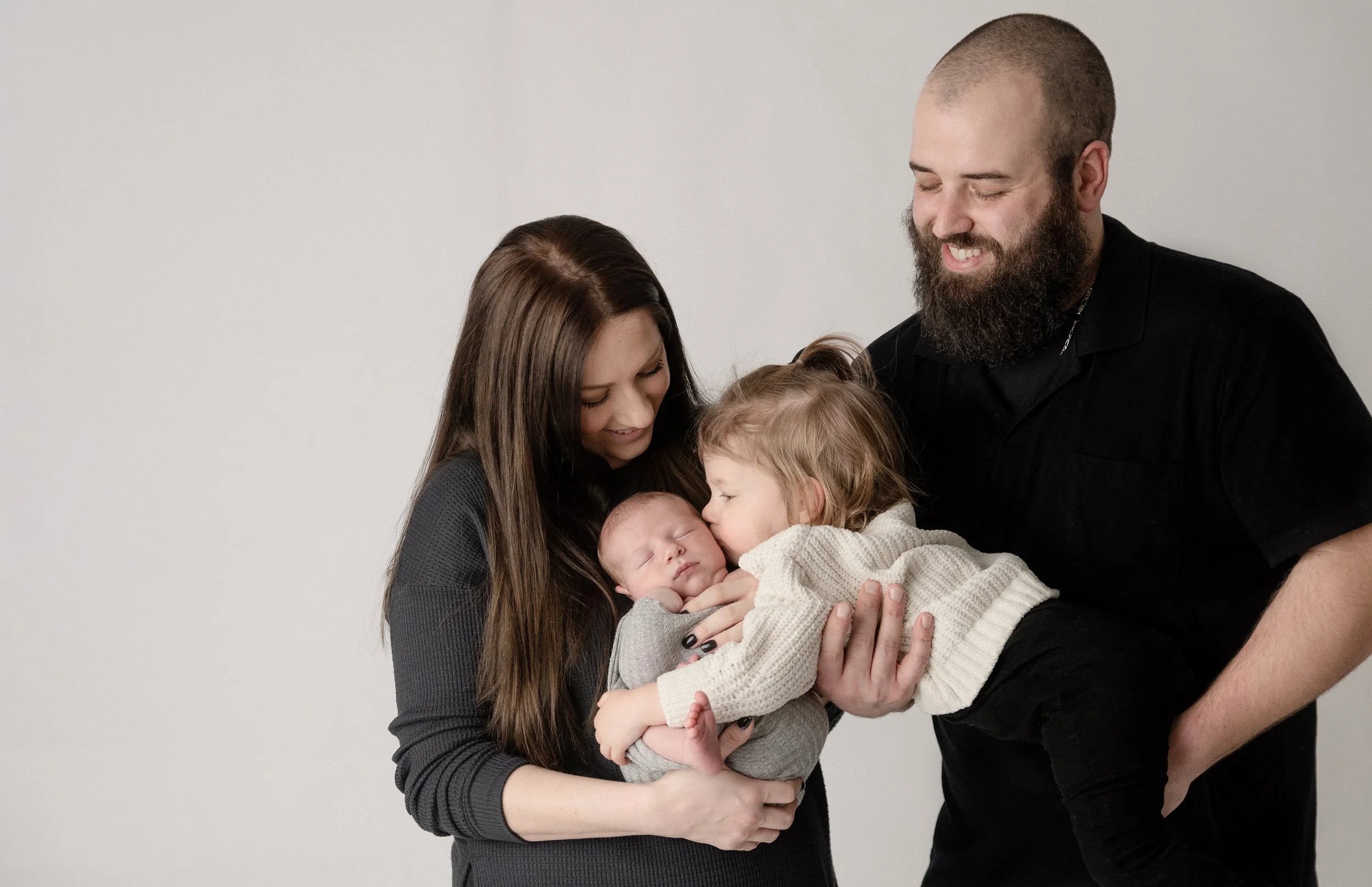 A woman and man hold two children, with the woman kissing a sleeping baby and the older child giving a kiss to the baby's forehead on a plain background.