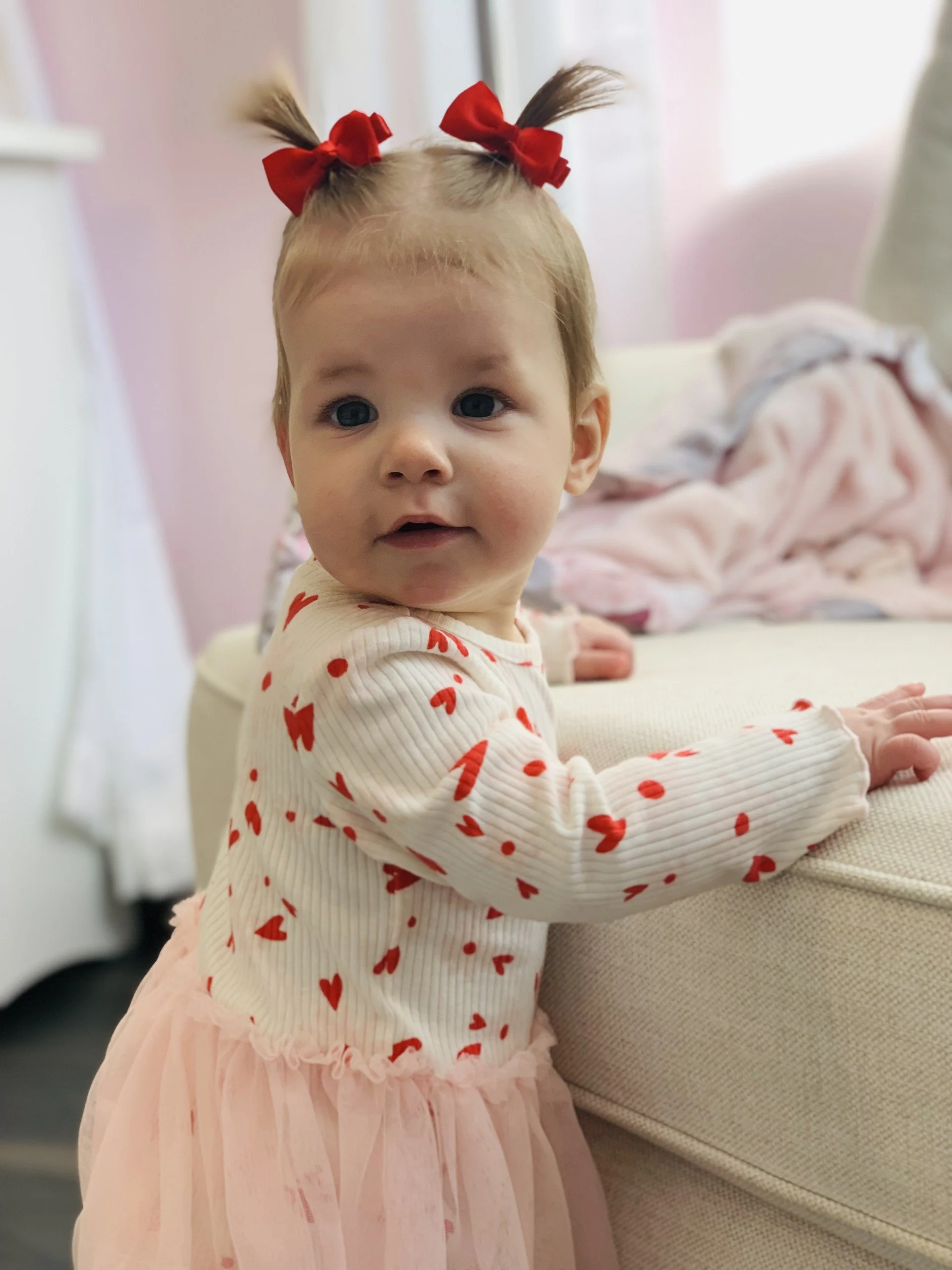 A young girl with light hair styled in pigtails secured with red bows, wearing a cream-colored top with red hearts and a pink tulle skirt, standing next to a beige sofa and looking at the camera.