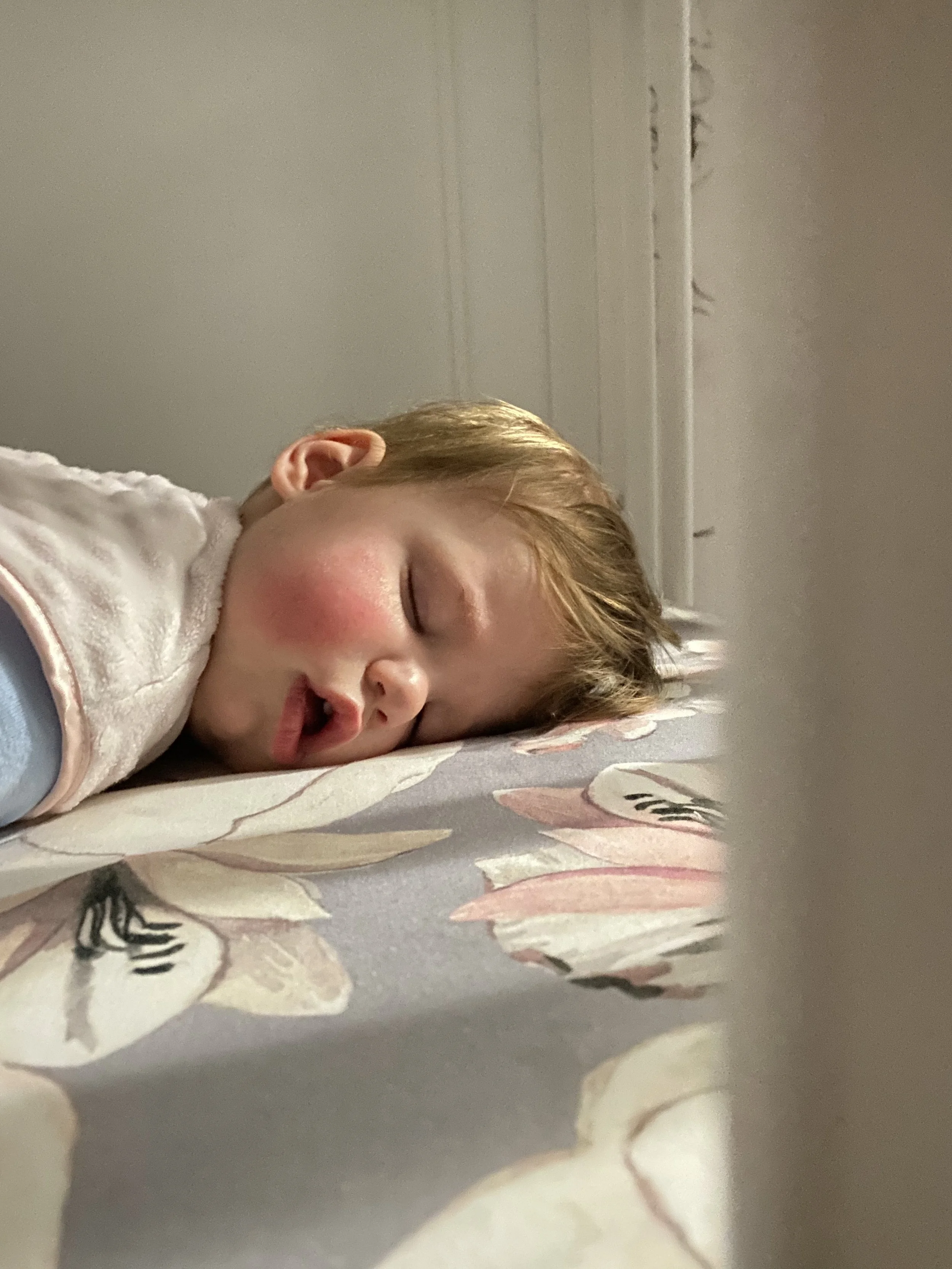 A young child with light brown hair sleeping on a floral bedspread in a bedroom.