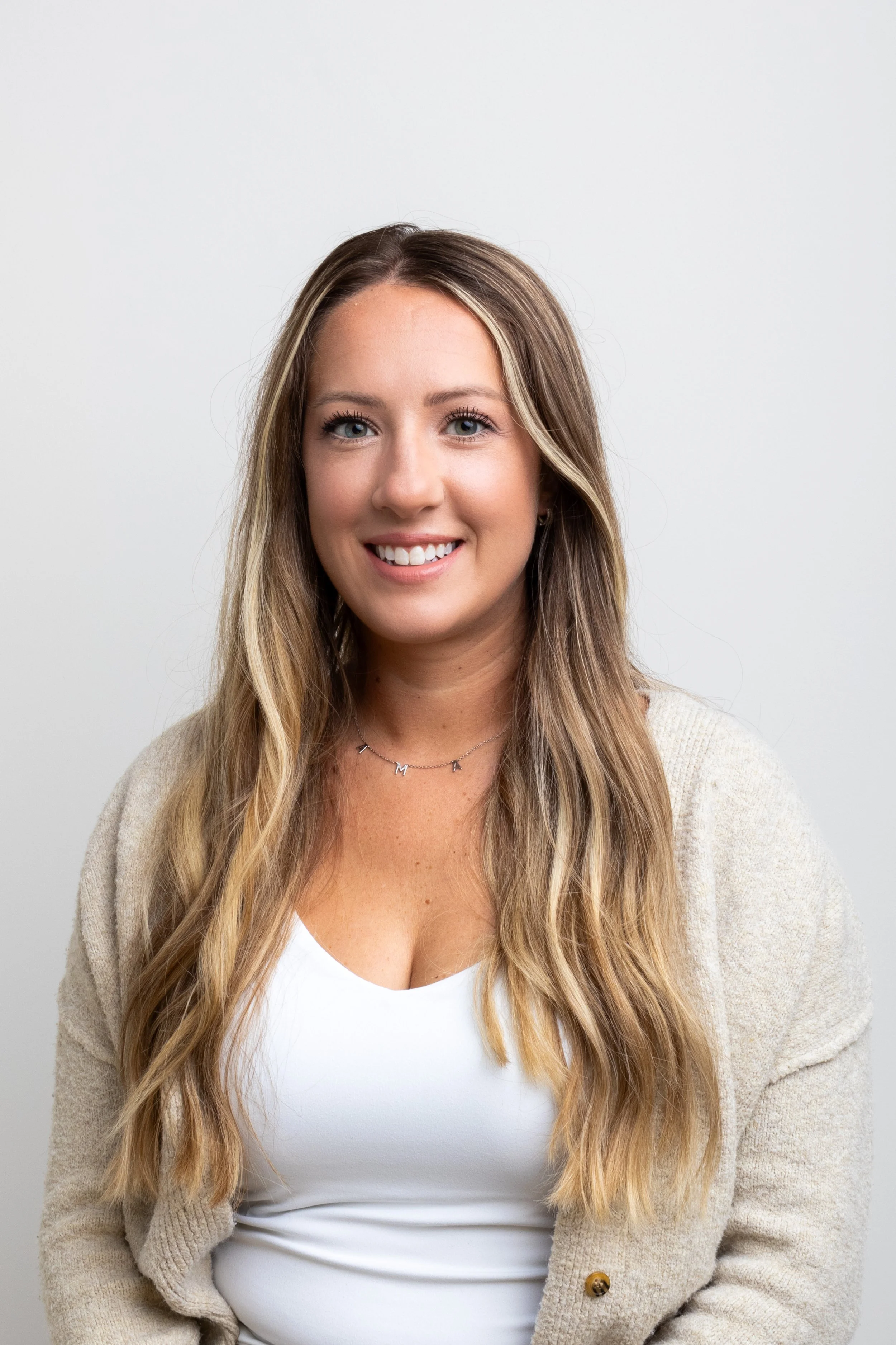 A woman with long wavy blonde hair, blue eyes, smiling, wearing a white top, a beige cardigan, and a delicate necklace, against a plain white background.
