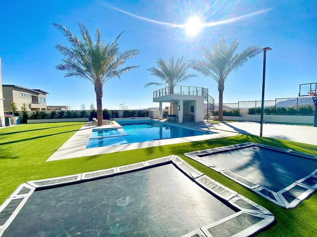 A bright backyard with a swimming pool, two palm trees, and two trampolines on a sunny day.