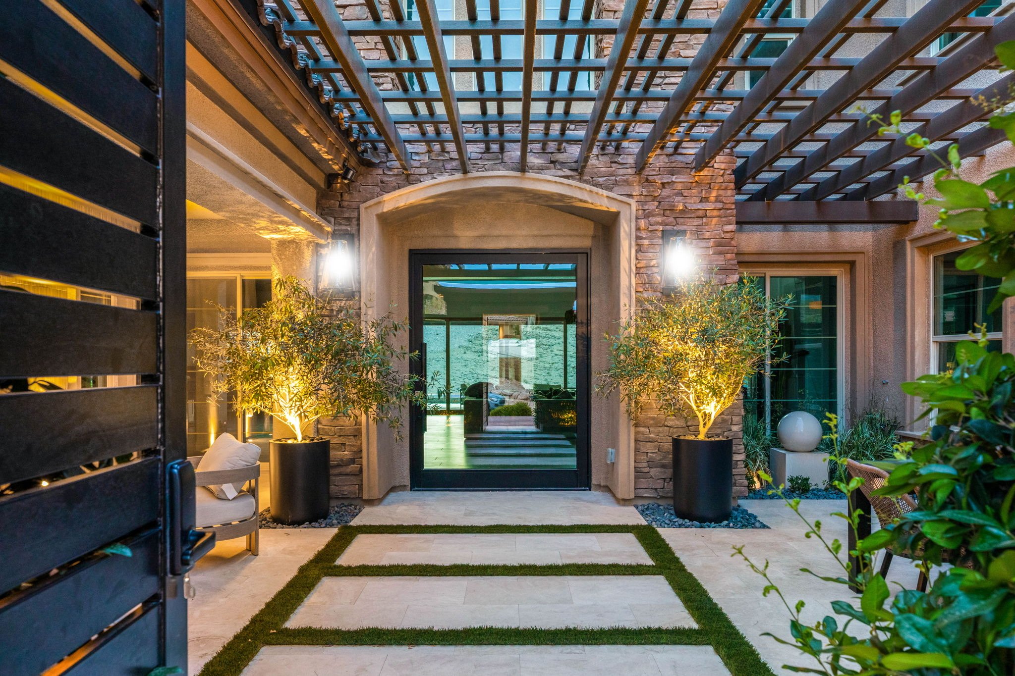 courtyard landscape design featuring custom wood pergola, stone cladding accents, potted olive trees, and paver stepping pads set in synthetic turf on Olympia Outlook Dr, Las Vegas.