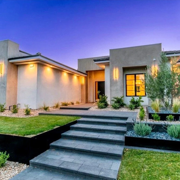 Modern house with a minimalist landscape, illuminated outside lighting, gray concrete steps leading to the entrance, and a well-manicured lawn and plants.