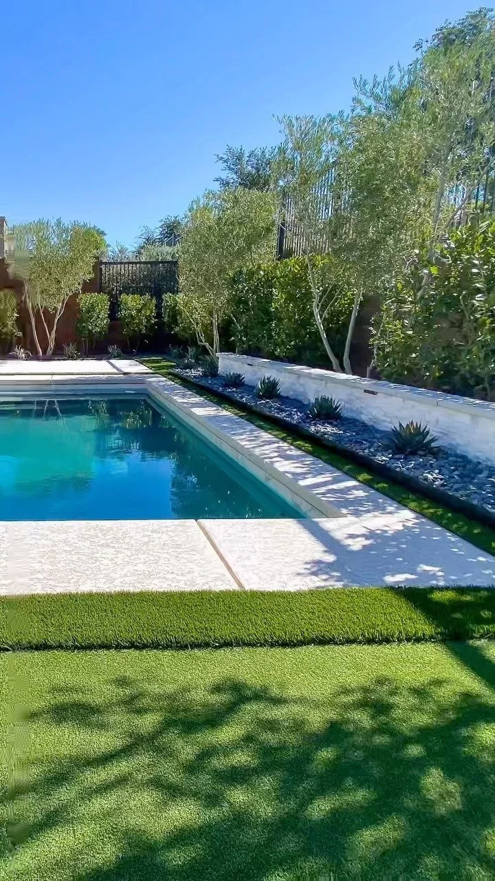 A backyard with a swimming pool, green grass, trees, and a high fence under a clear blue sky.