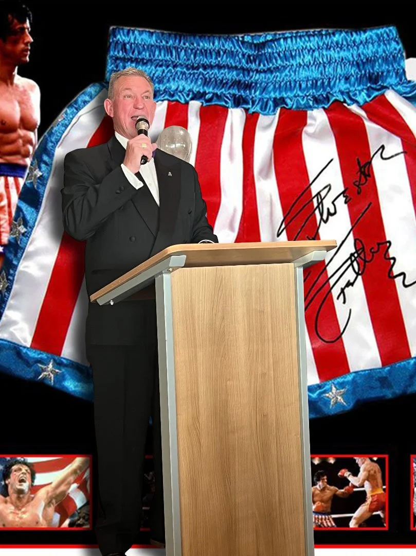 A man in a tuxedo speaking at a podium in front of a large background image of boxing shorts with red and white stripes and a blue waistband, signed with a black marker. The background also includes images of boxers in action, with one shirtless boxer celebrating and another two boxing.