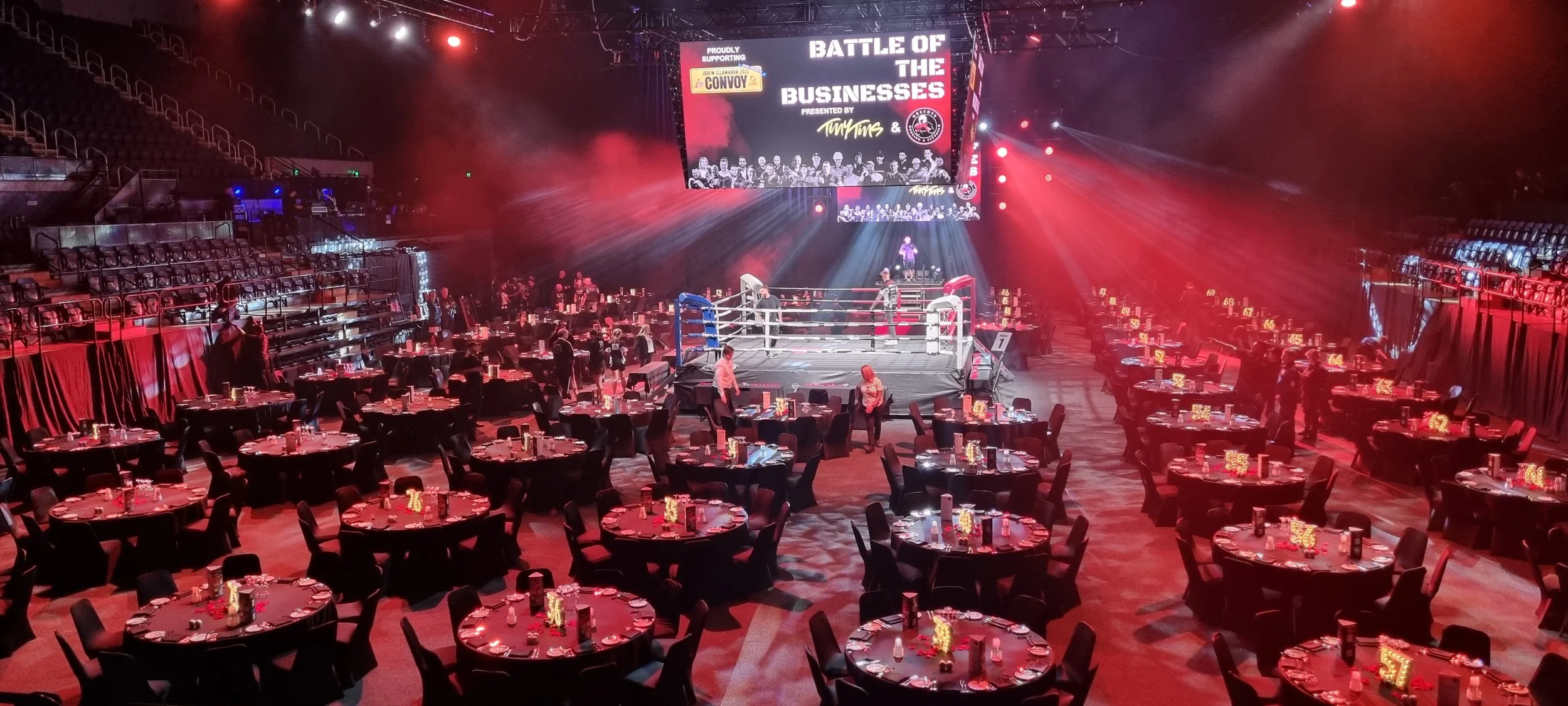 Empty boxing ring in a large event hall with round tables set for dining, dim lighting, and a large screen displaying 'Battle of the Businesses'