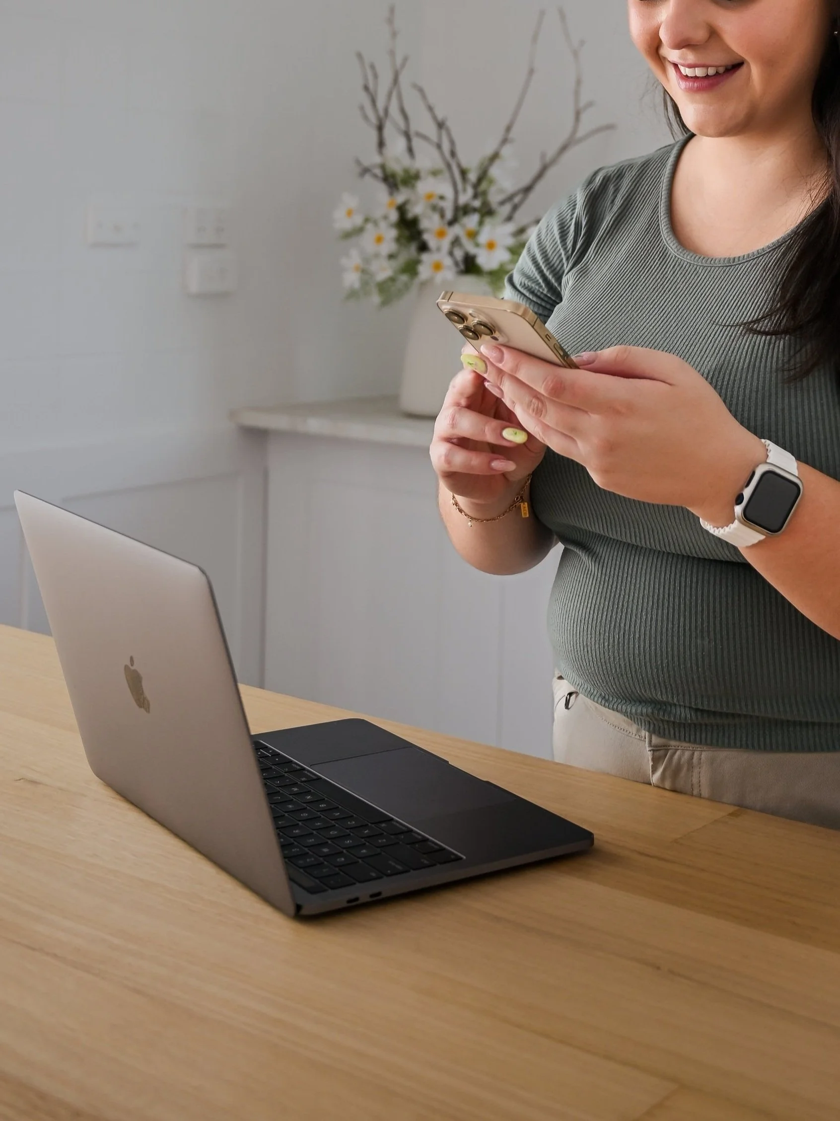 A woman standing at a wooden table using a smartphone, with a closed laptop nearby and a vase with flowers and branches in the background.