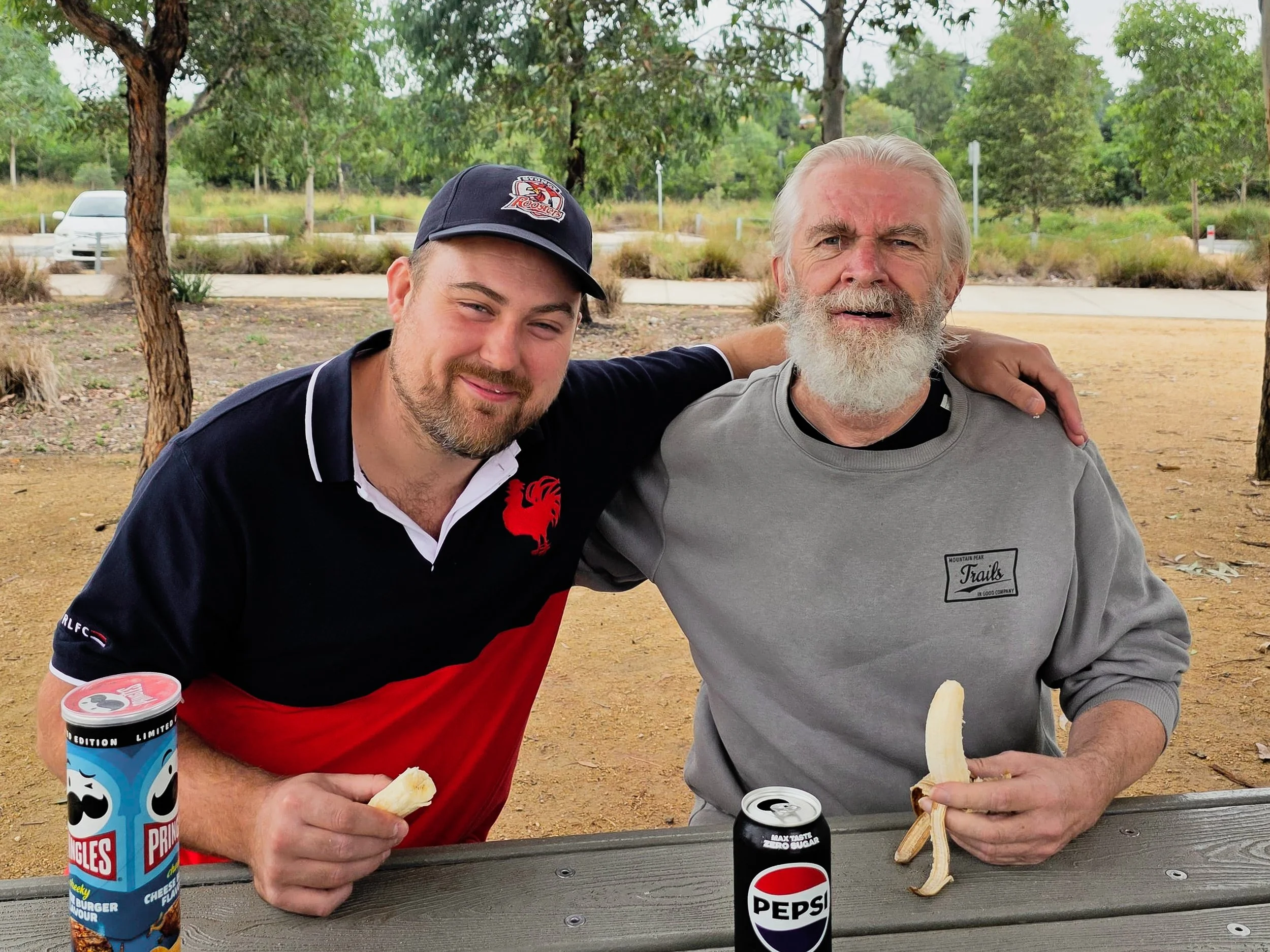 Two men sitting at a picnic table outdoors, each eating a banana, with snacks and drinks including a Pringles can and a Pepsi can in front of them. One man has a beard and gray hair, wearing a gray sweatshirt, and the other has a shorter beard, weari
