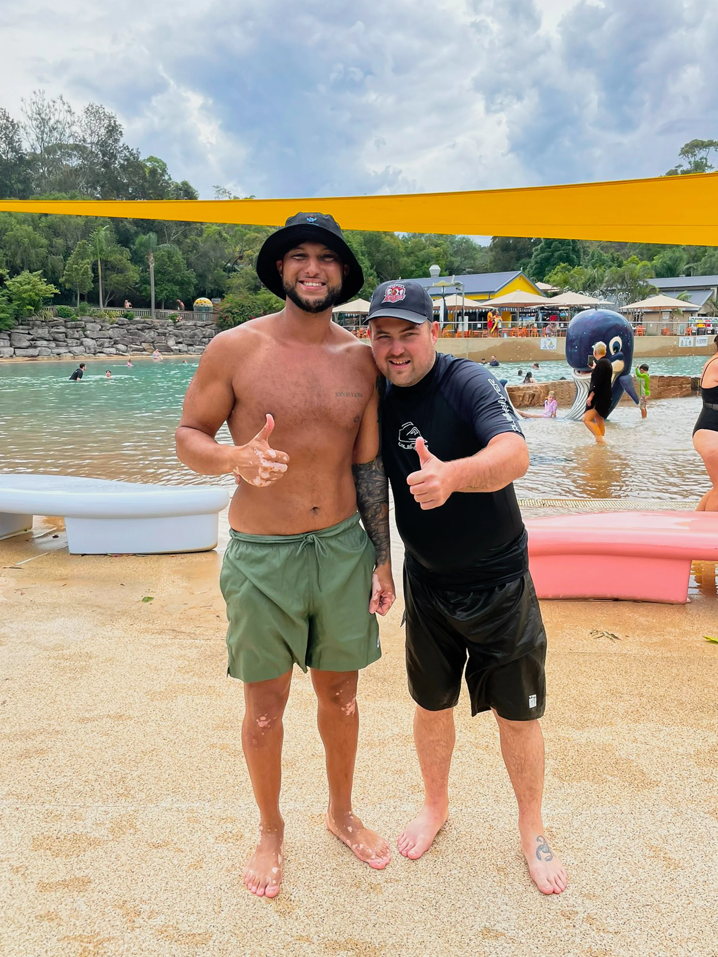 Two men standing on a sandy beach at a water park, smiling and giving thumbs-up. One shirtless with a hat and the other in a black shirt and black shorts. Water and other visitors visible in the background.