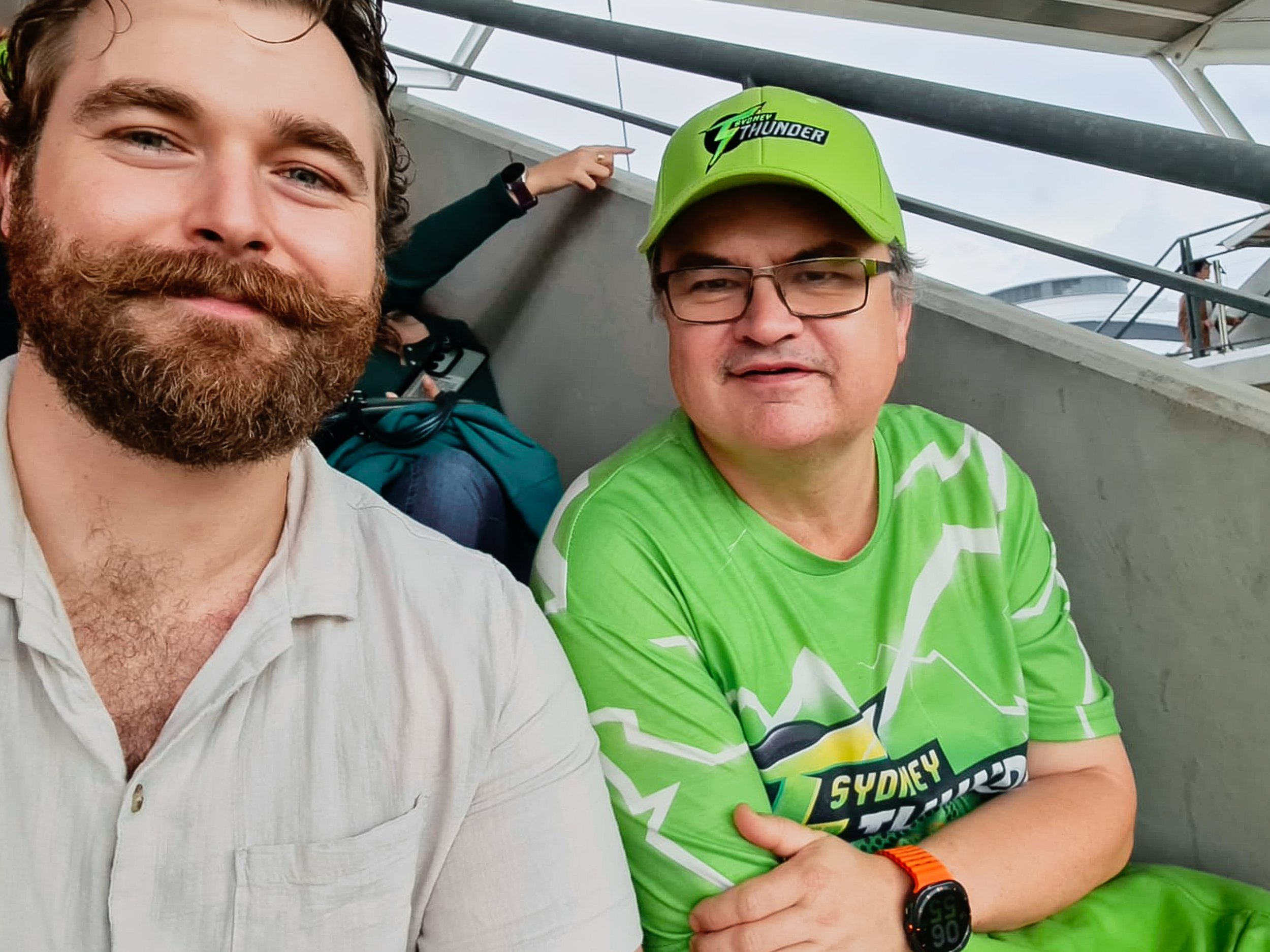Two men sitting next to each other on a stadium or arena seating section. The man on the left has a beard and is smiling, and the man on the right is wearing glasses, a bright green Sydney Thunder cap, and a matching sports shirt. The background show
