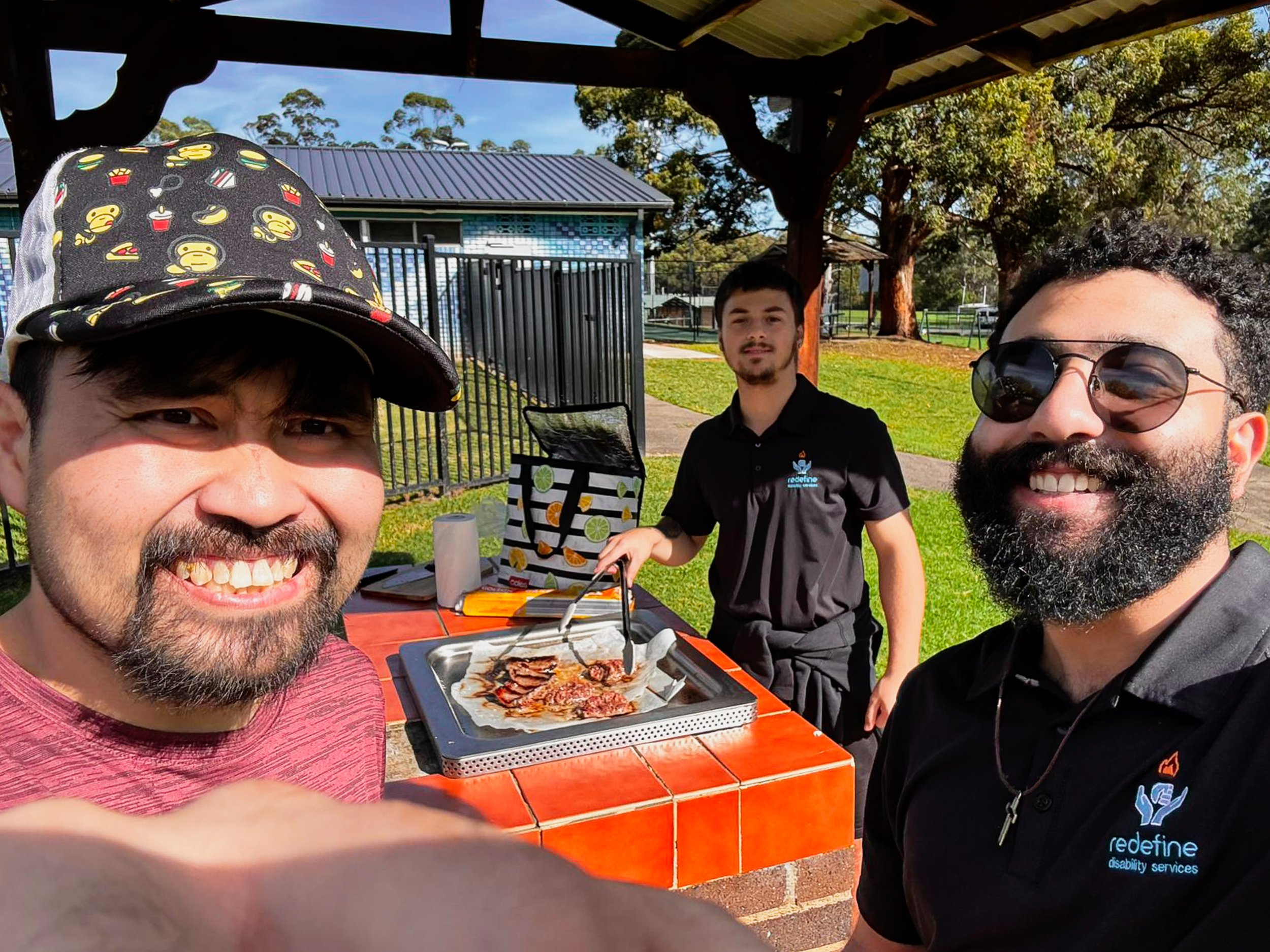 Three men smiling outdoors at a barbecue grill, with one cooking meat. One man wears a baseball cap with cartoon monkey faces. The other two wear black shirts with 'redefine disability services' logo. Background includes trees, a fence, and a buildin