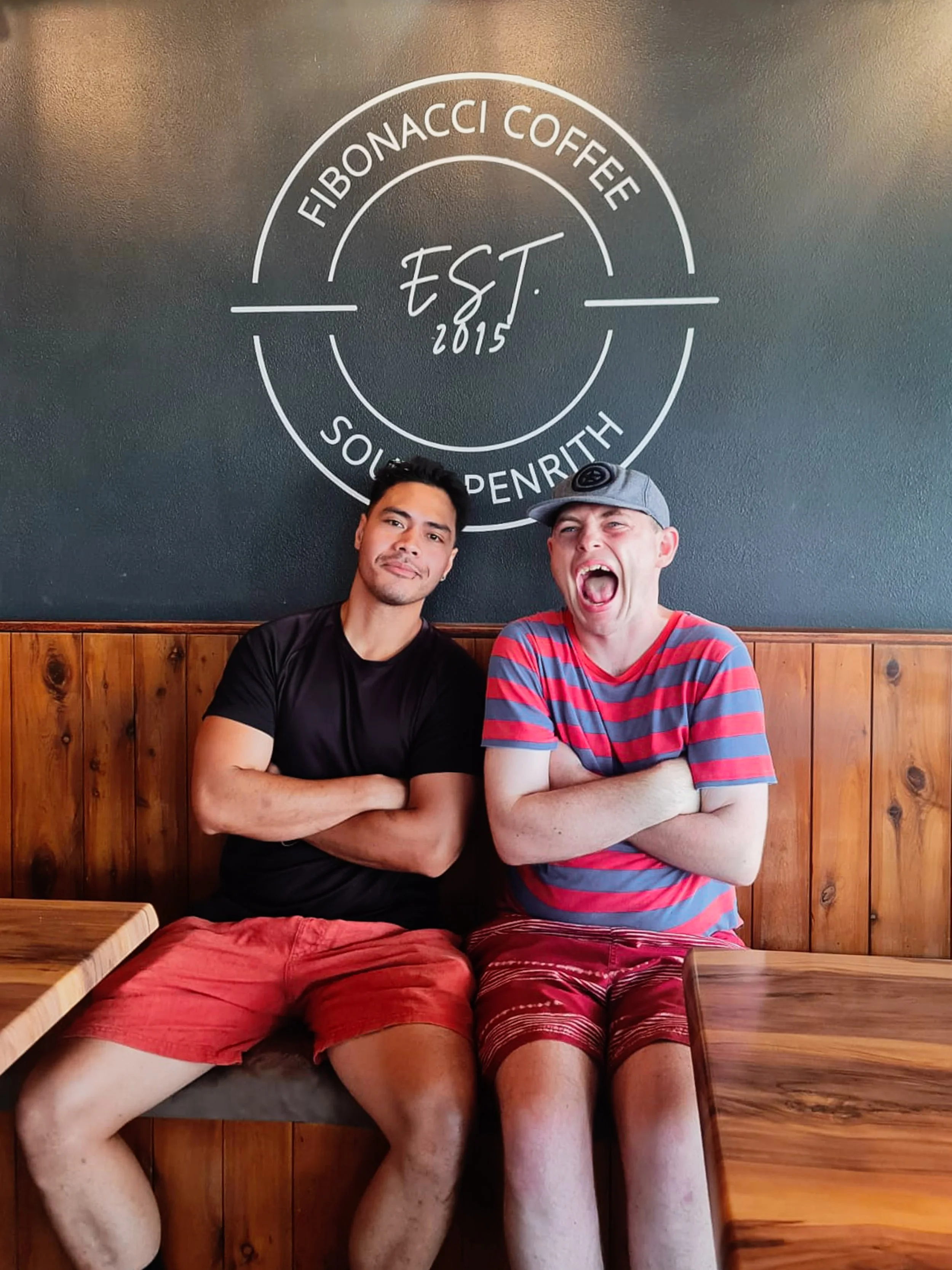 Two men sitting in a coffee shop with a dark green wall behind them featuring a white logo that reads Fibonacacci Coffee South Penrith. The man on the left is wearing a black t-shirt and red shorts, with his arms crossed. The man on the right is wear