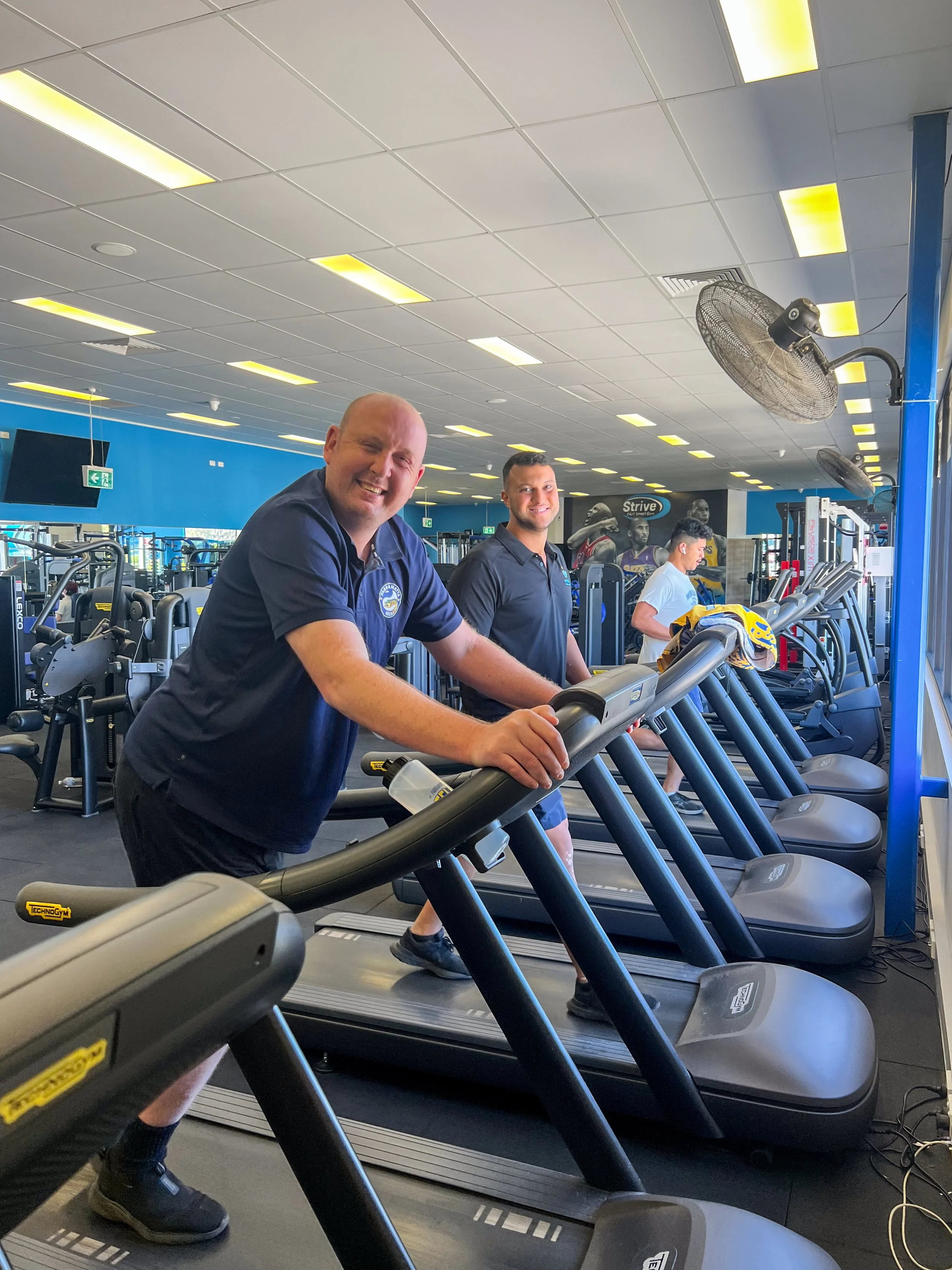 Three smiling men in navy shirts exercise on treadmills in a spacious gym with bright lighting and blue walls.
